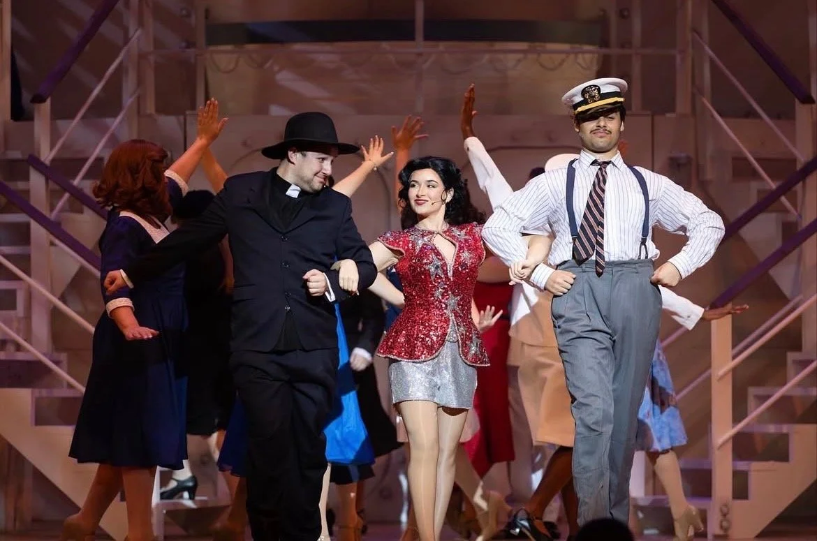 Actors in costumes performing a musical or dance on stage with a wooden staircase in the background.