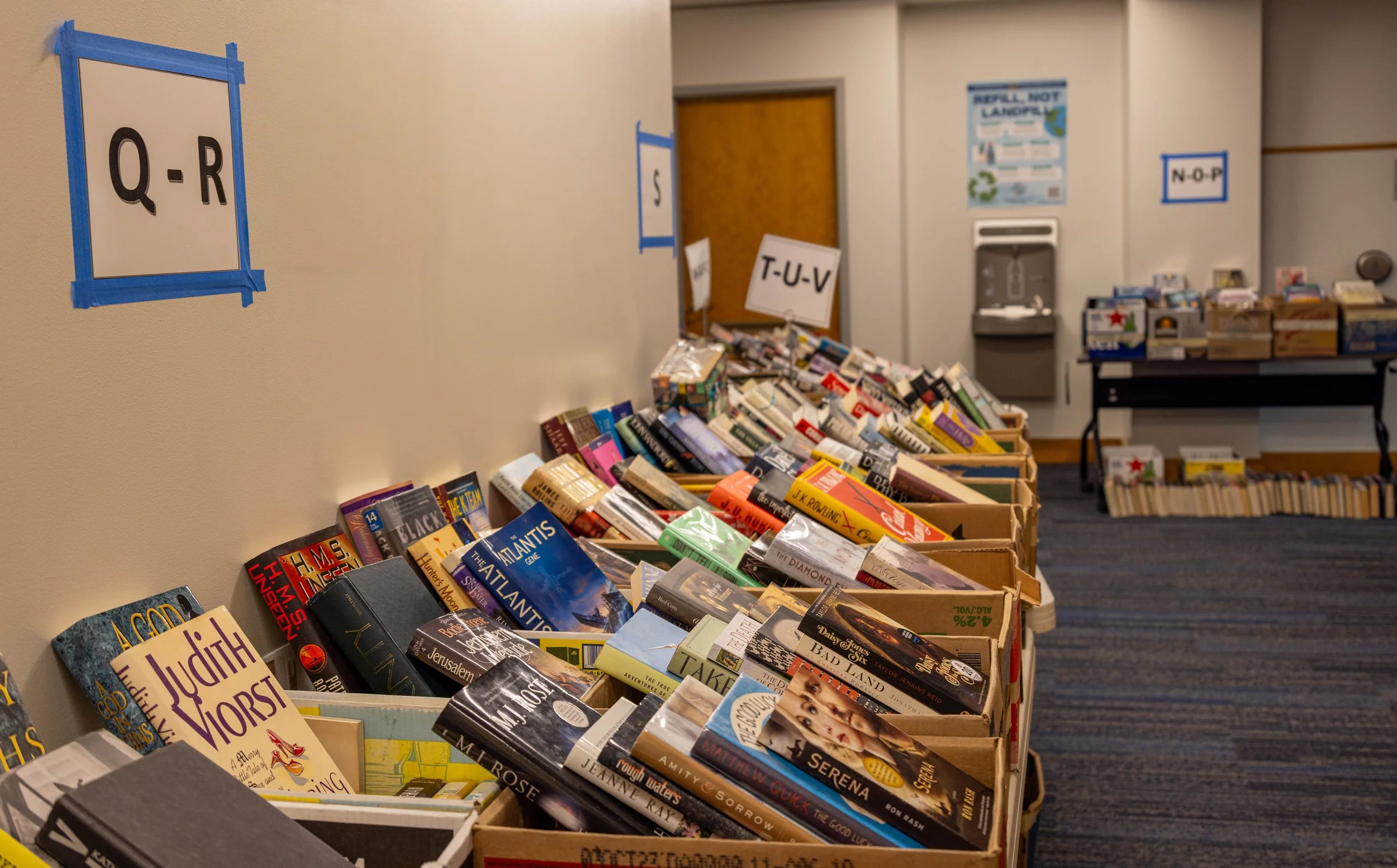 Books organized by alphabet in bins and on tables in a library or bookstore, with signs indicating the alphabetical sections.