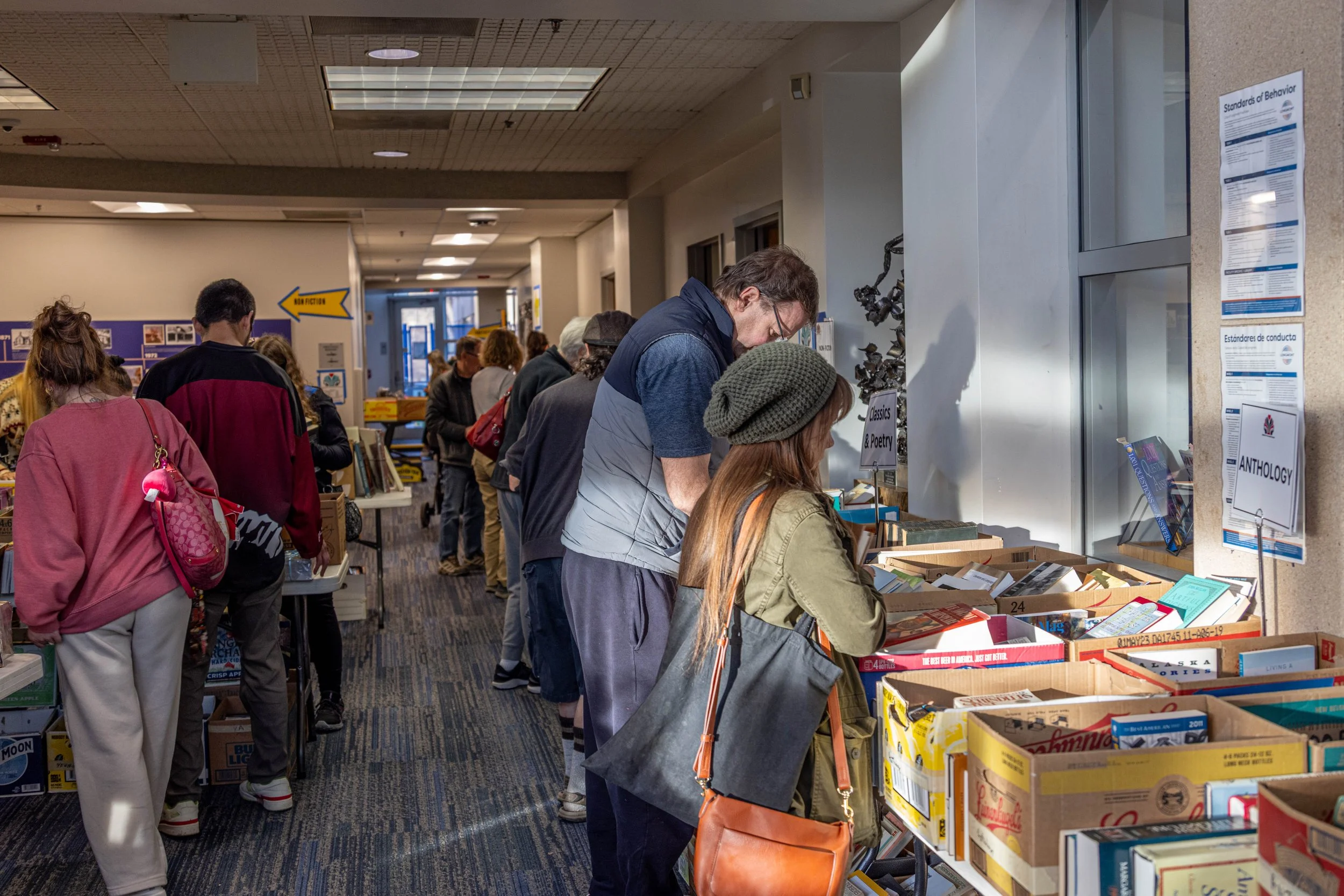 People browsing books at a sale or bookstore with boxes of books on tables and shelves in the background.