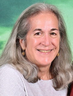 A woman with long gray hair smiling outdoors with lush green trees in the background.