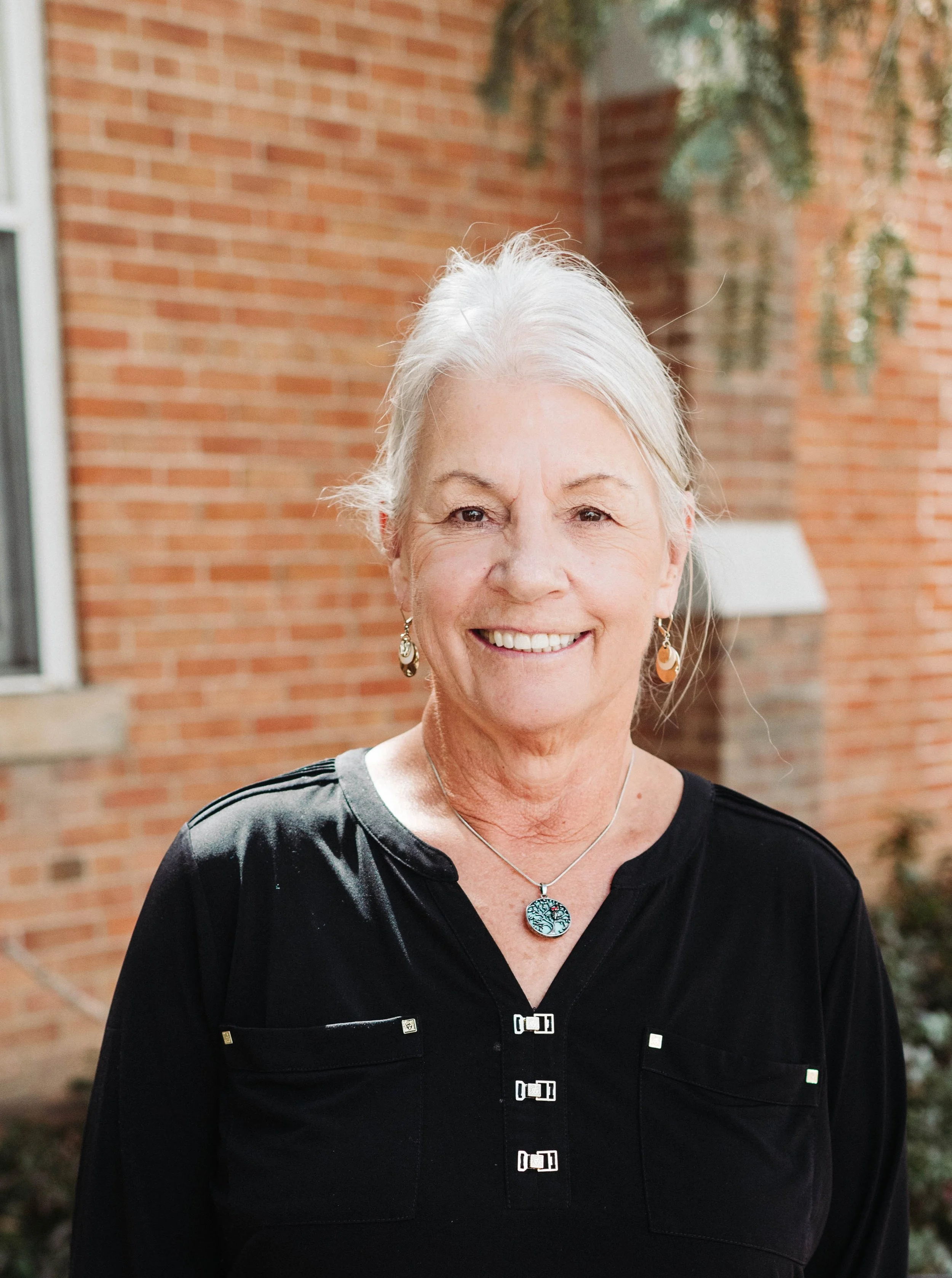 An elderly woman with white hair smiling outdoors in front of a brick building, wearing a black blouse, earrings, and a necklace with a tree pendant.