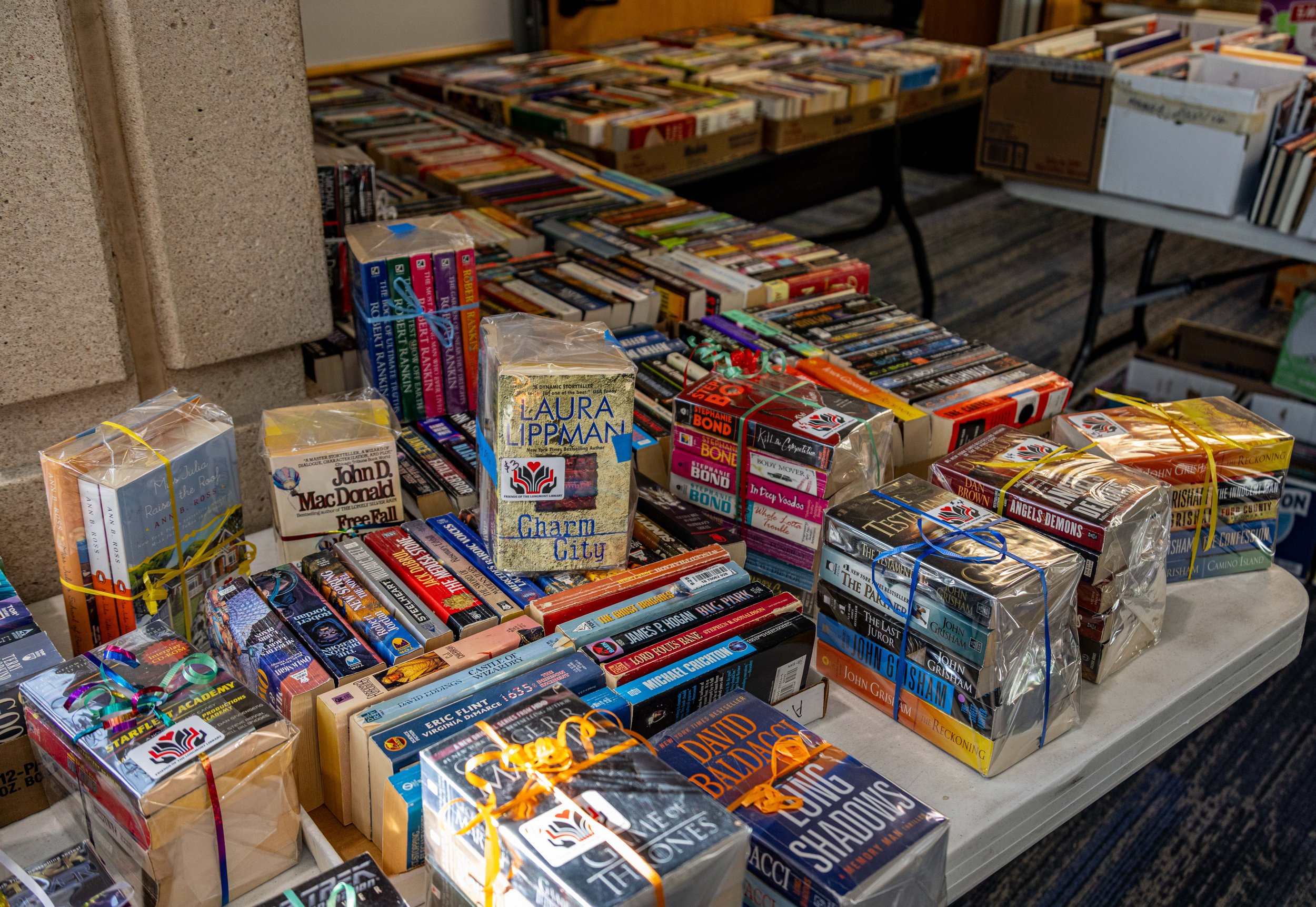Tables of stacked books at a book sale, with some wrapped in plastic and tied with colorful ribbons.