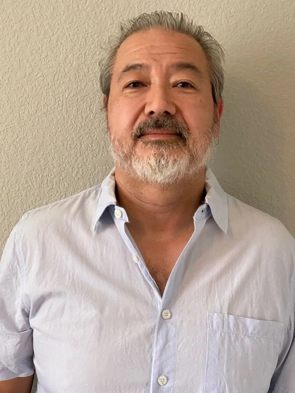 A middle-aged man with gray hair and a beard, wearing a light gray button-up shirt, standing against a textured beige wall.