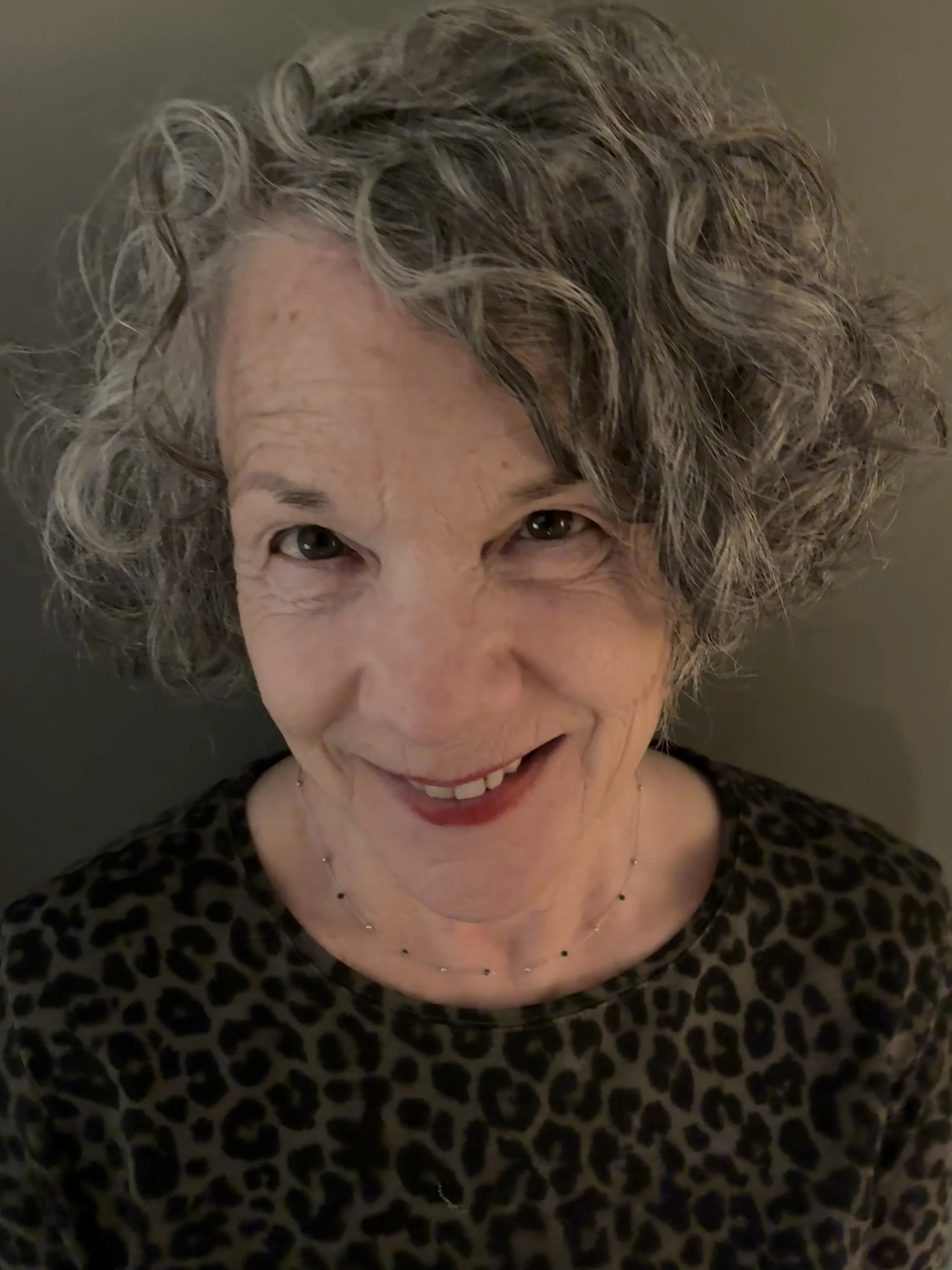 A smiling elderly woman with curly gray hair, wearing a leopard print top and a delicate necklace.