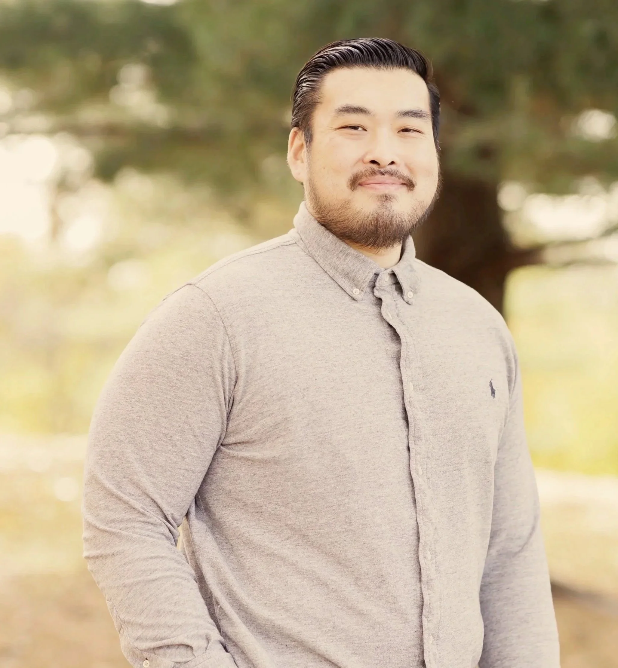 A man with dark hair and a beard standing outdoors in front of a blurred background of trees, wearing a light gray button-up shirt with a small logo on the chest.