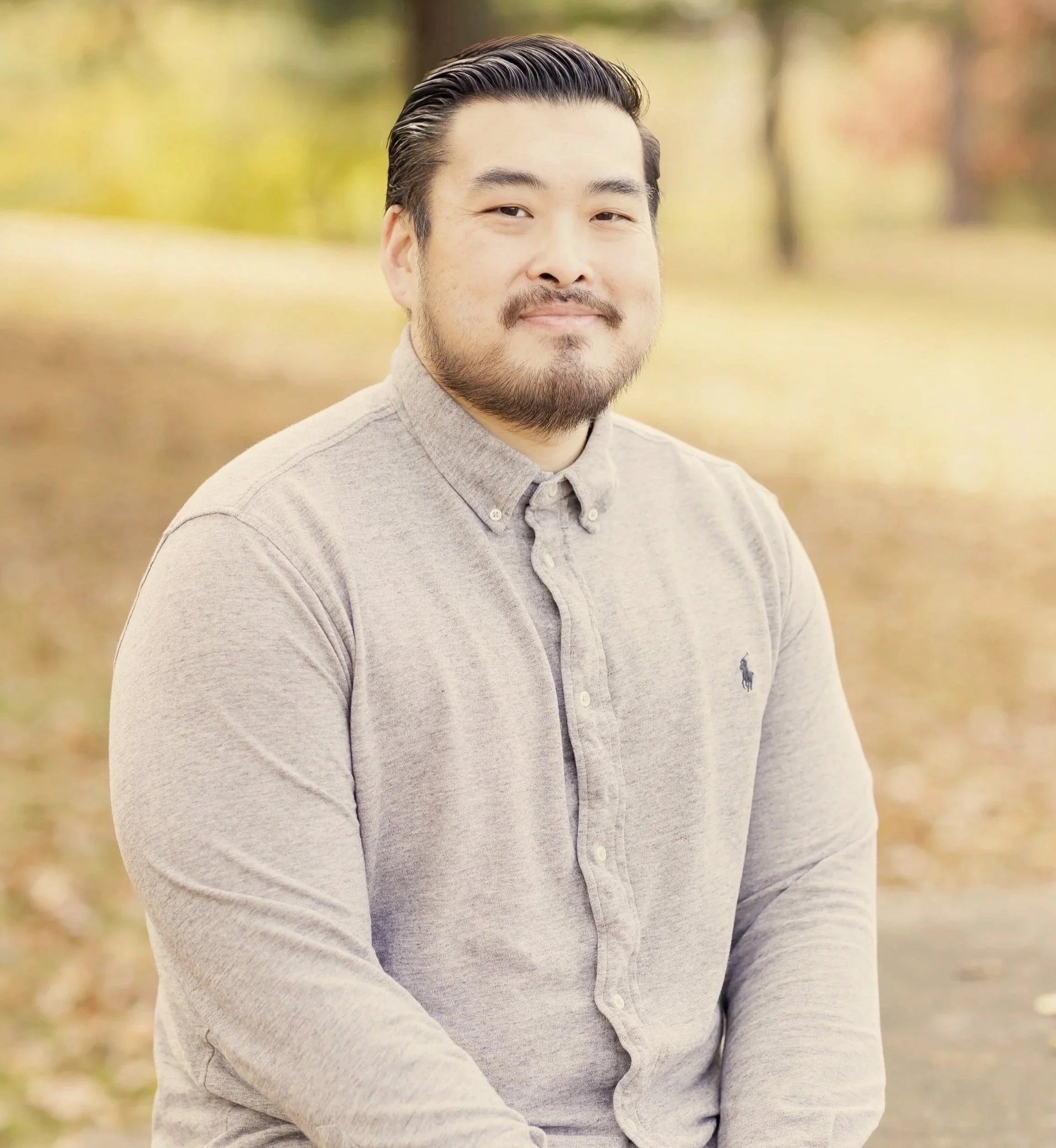 A man with a beard and slicked-back hair, wearing a light gray button-up shirt, standing outdoors with blurred autumn trees and fallen leaves in the background.