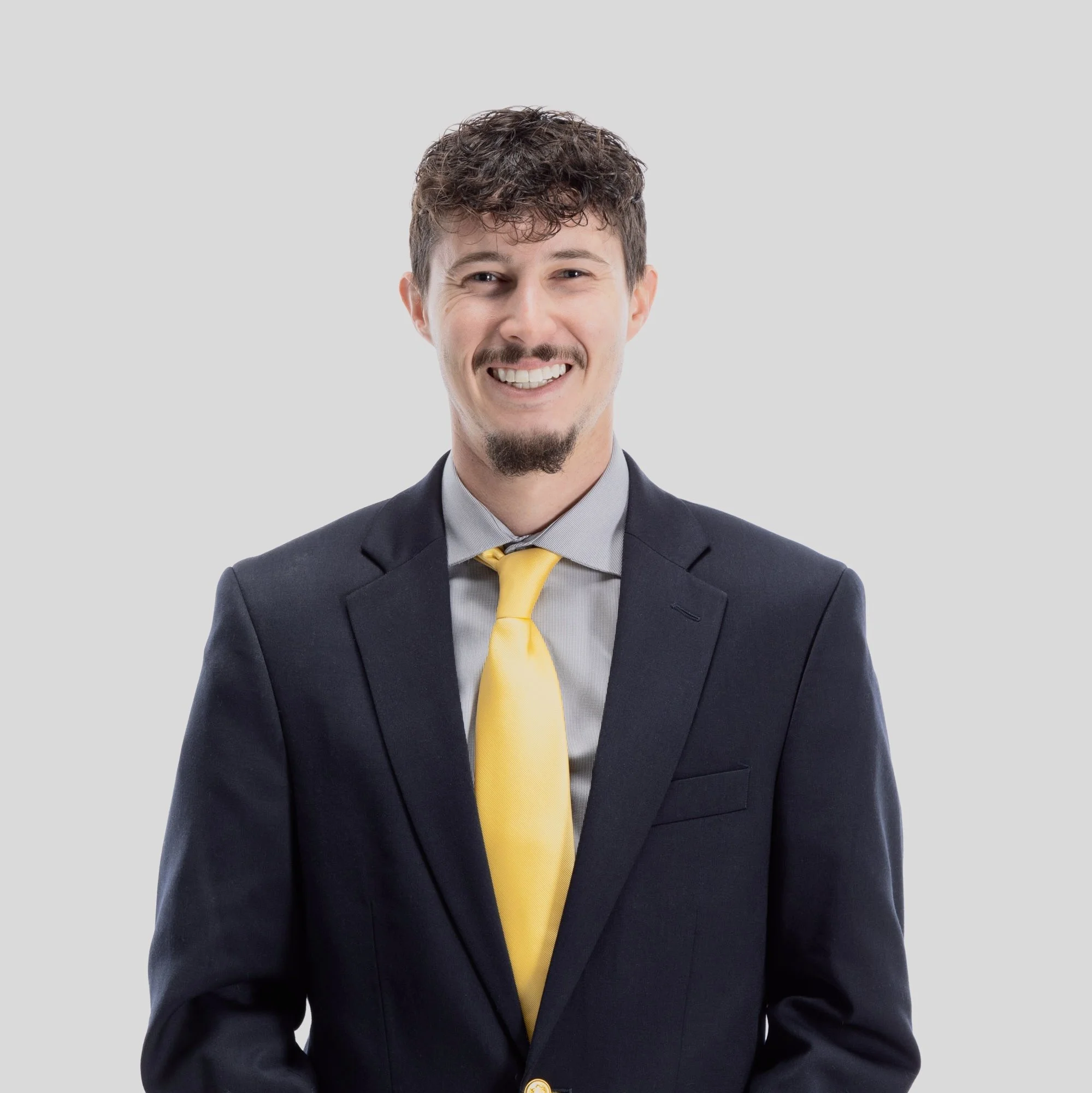 A smiling man with short curly hair, light facial hair, wearing a dark suit, gray shirt, and yellow tie, standing against a plain light gray background.
