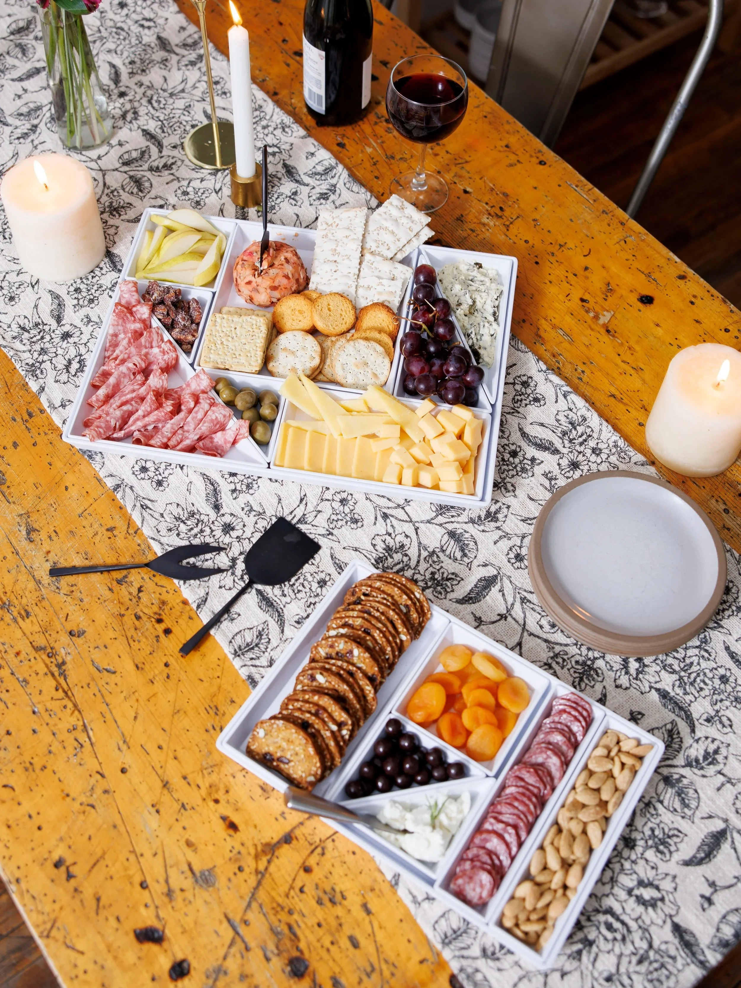 A cheese and charcuterie platter with grapes, crackers, sliced meats, cheeses, nuts, and dried fruit, on a rustic wooden table with candles and a wine glass nearby.