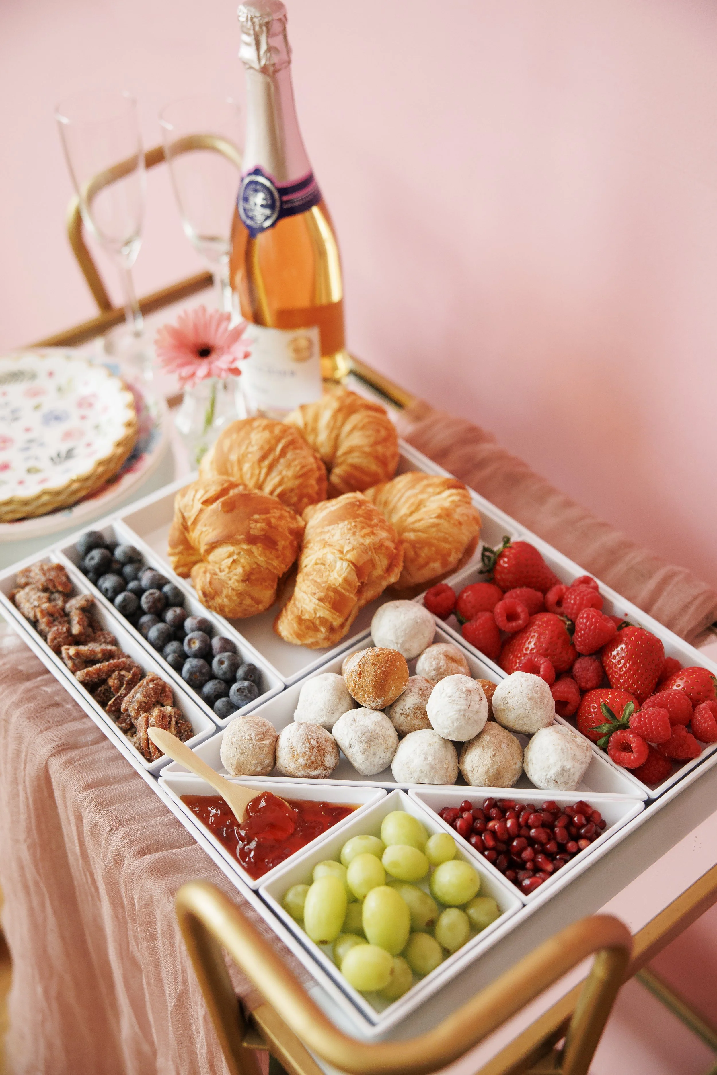 A tray with croissants, strawberries, raspberries, blueberries, powdered donuts, and various jams and candies on a pink table against a pink wall.