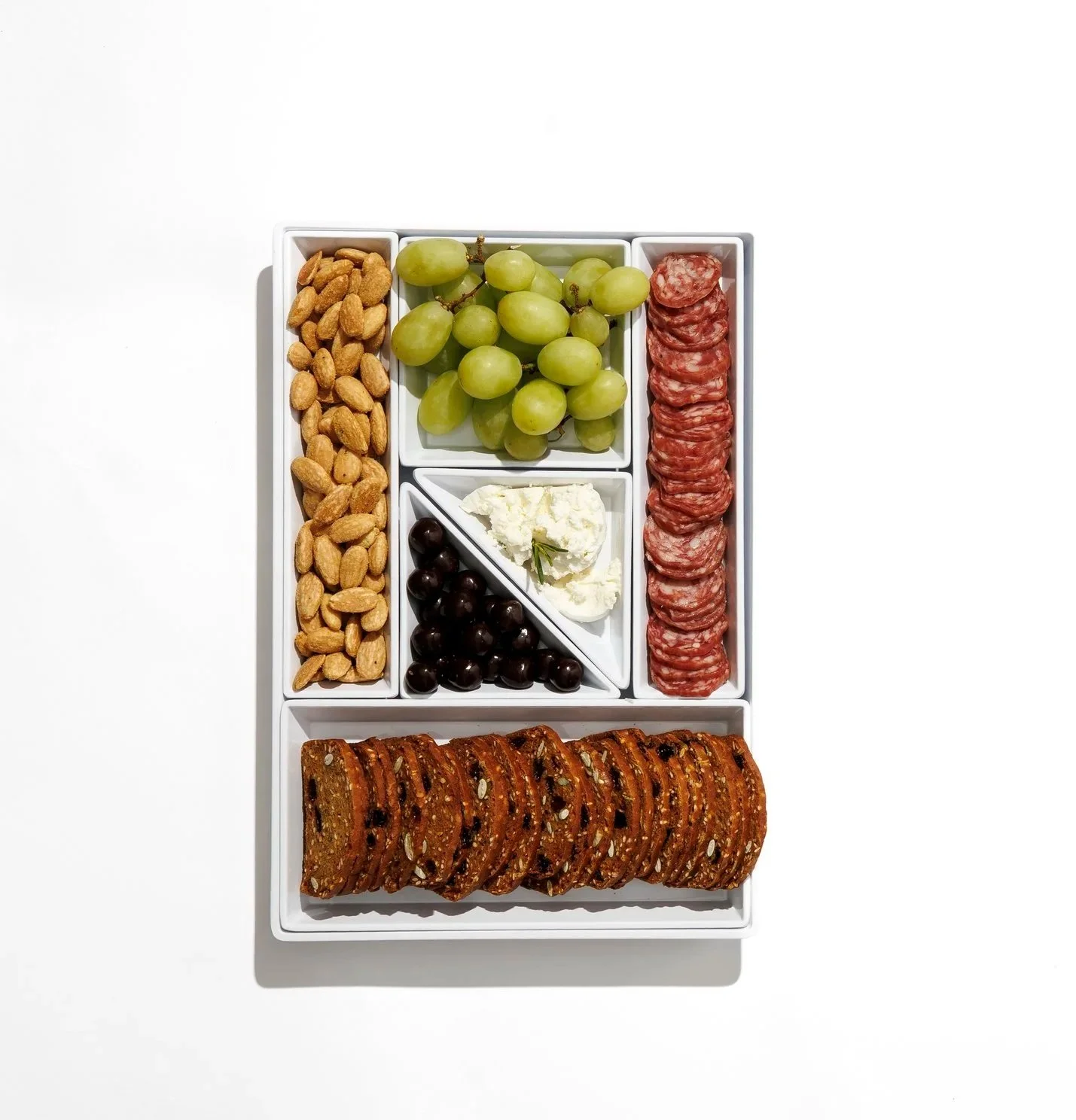 Charcuterie board with green grapes, sliced salami, black olives, feta cheese, roasted nuts, and thinly sliced crisp bread.