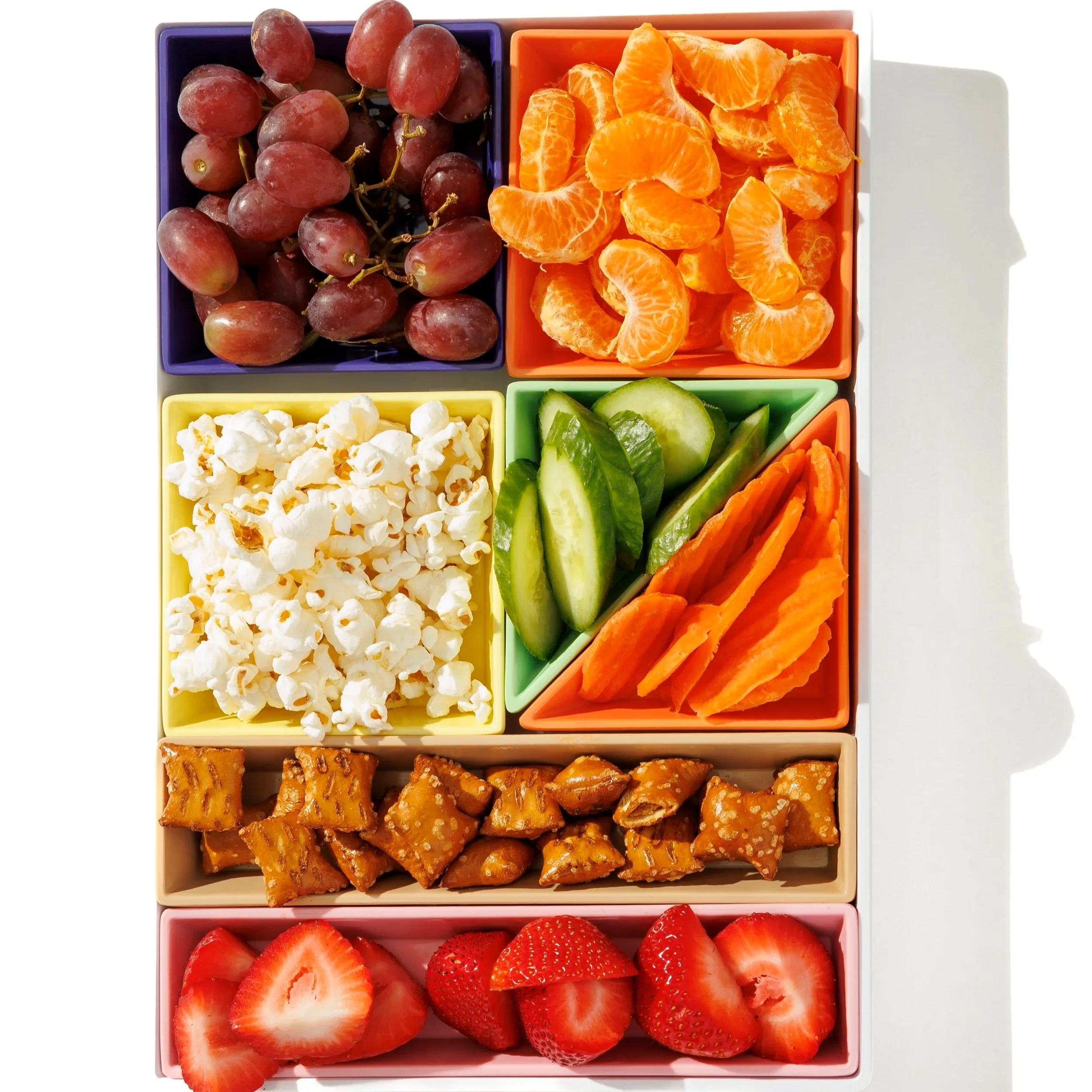 A colorful bento box with compartments containing grapes, orange slices, strawberries, chopped carrots, cucumber slices, popcorn, and piece of caramel brittle.