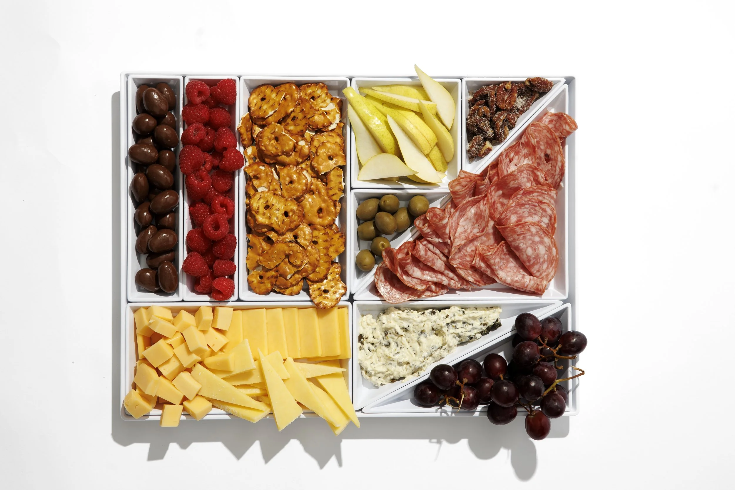 A white serving tray with small compartments holding assorted cheeses, fruits, and charcuterie. Visible items include cheddar cheese, blue cheese, grapes, raspberries, strawberries, pear slices, green olives, salami, and chocolate-covered nuts.