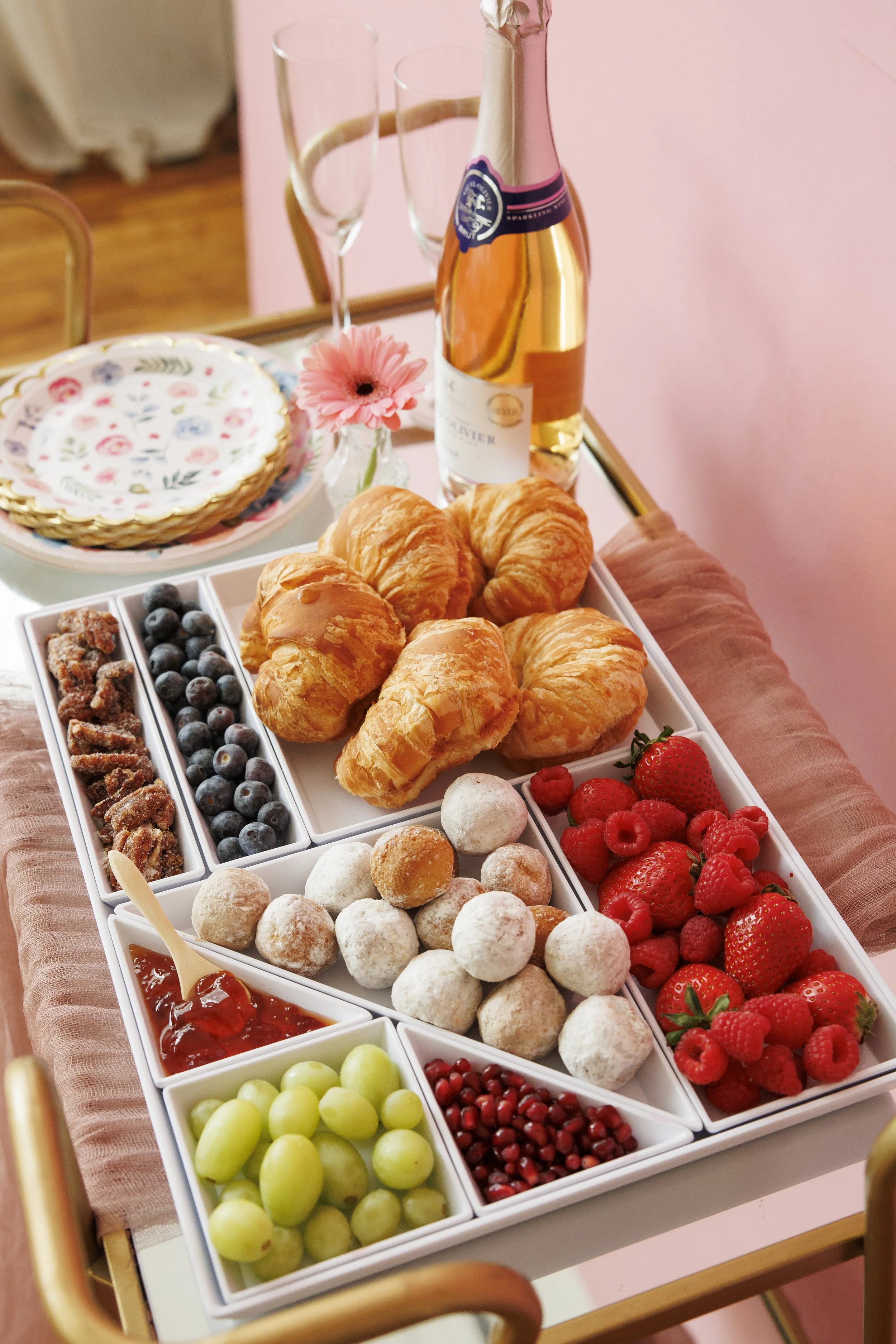 A serving tray with various fruits, pastries, and desserts, including croissants, strawberries, raspberries, blueberries, and assorted candies, with a bottle of sparkling wine and two empty champagne glasses in the background.