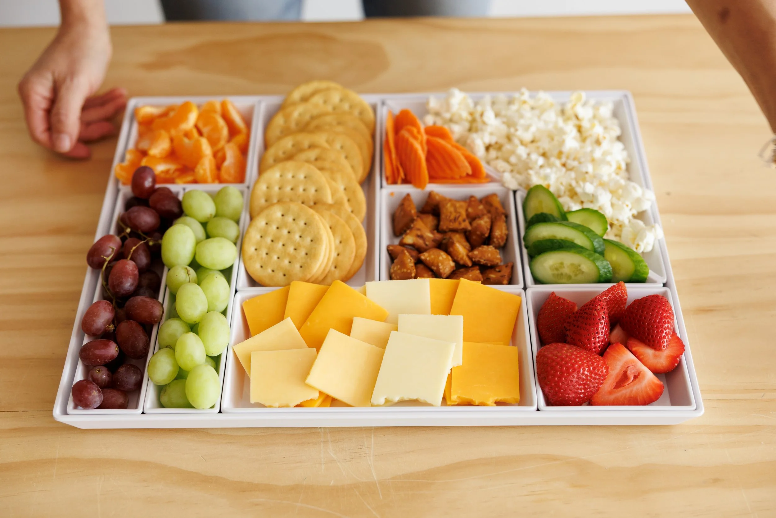 A white serving tray with various sections containing grapes, cheese, crackers, sliced vegetables, strawberries, and other snacks.