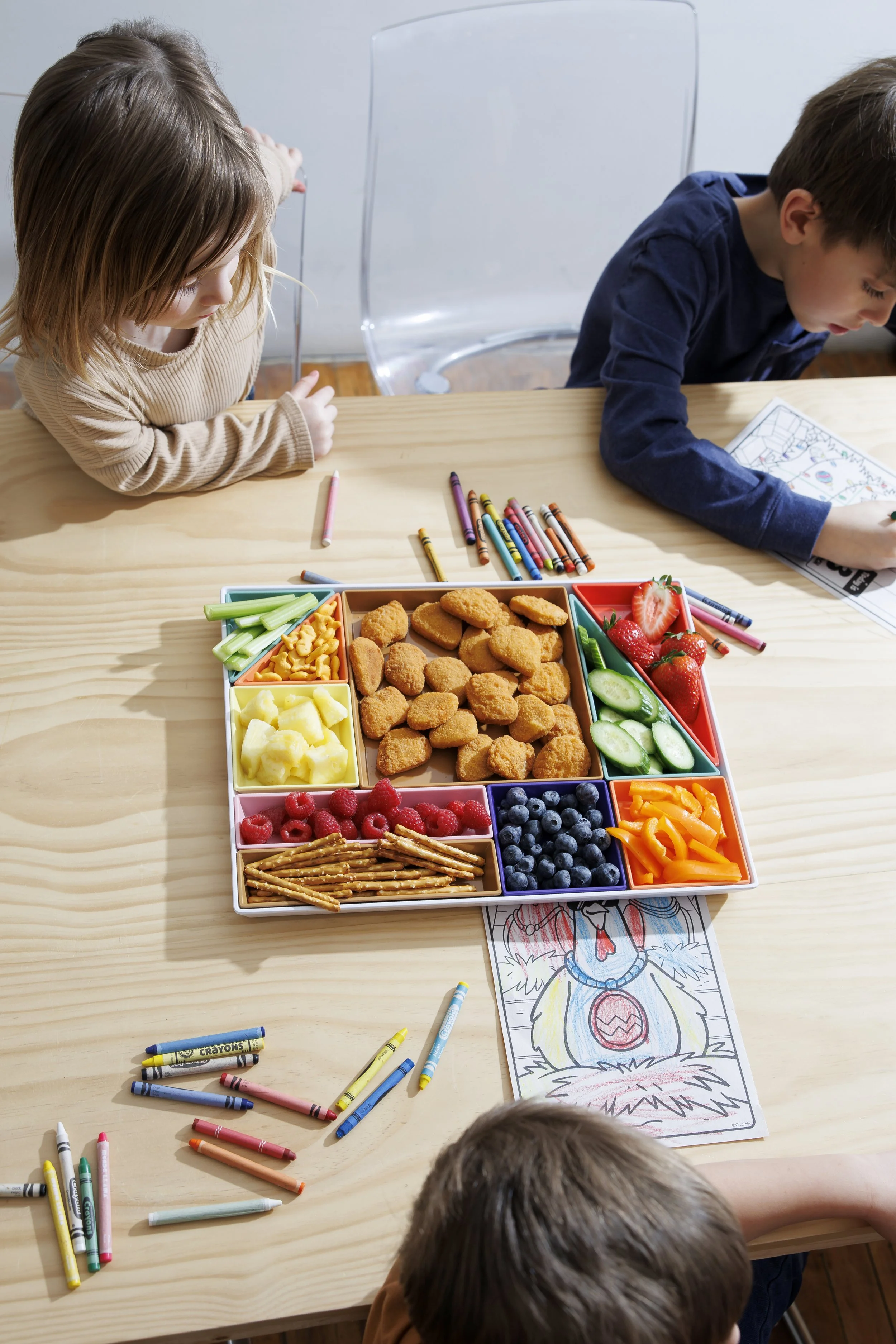 Children coloring a holiday-themed drawing on a table with crayons and a tray of snacks including fruit and crackers.