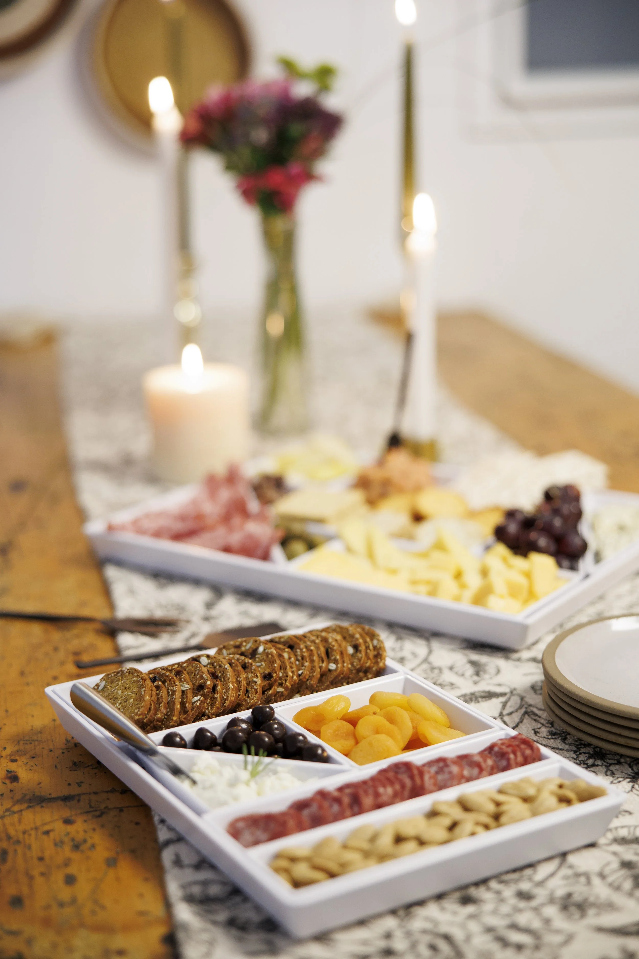 A cheese platter with pepperoni, grapes, and crackers, with a blurred background of a table with candles, flowers, and plates.