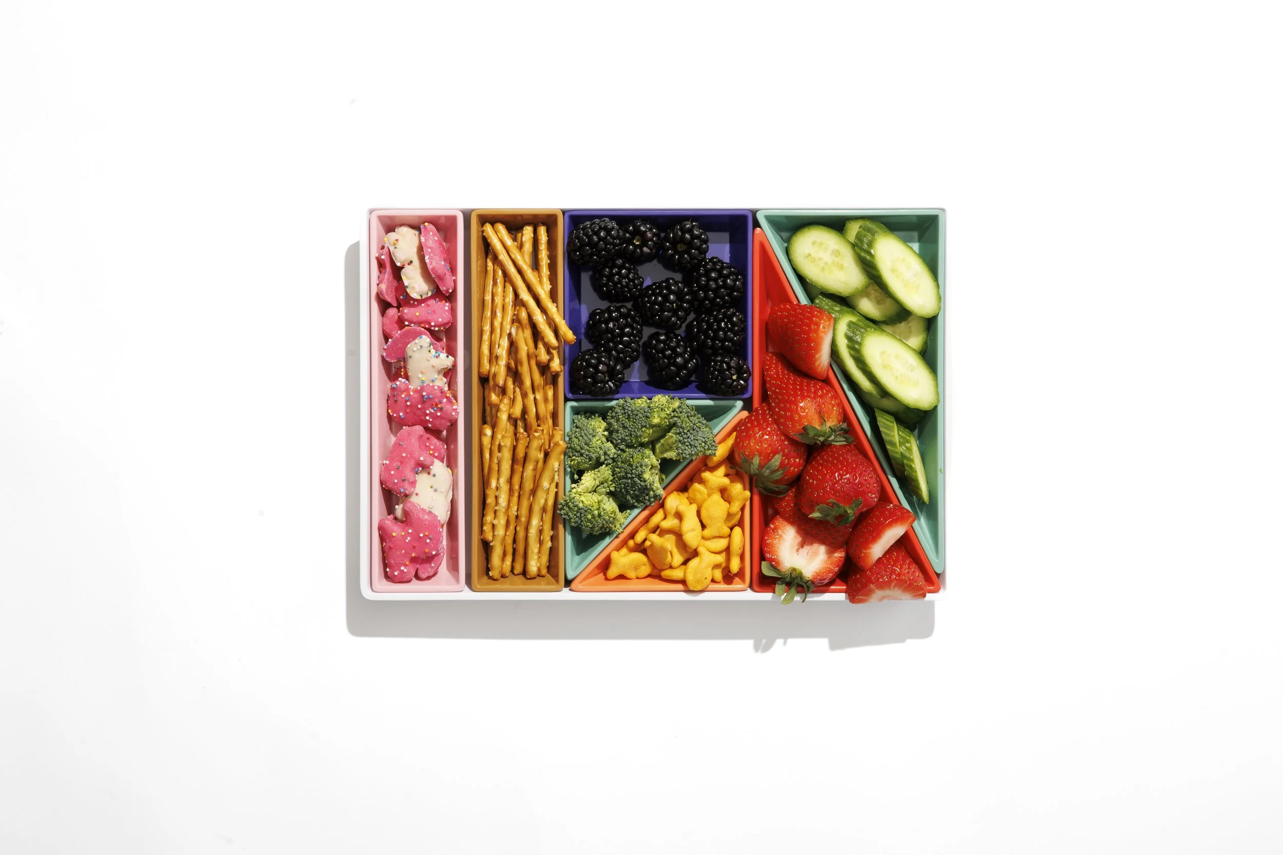 Colorful divided tray with pink animal-shaped cookies, pretzels, blackberries, sliced cucumbers, strawberries, broccoli, and cheese shapes on a white background.