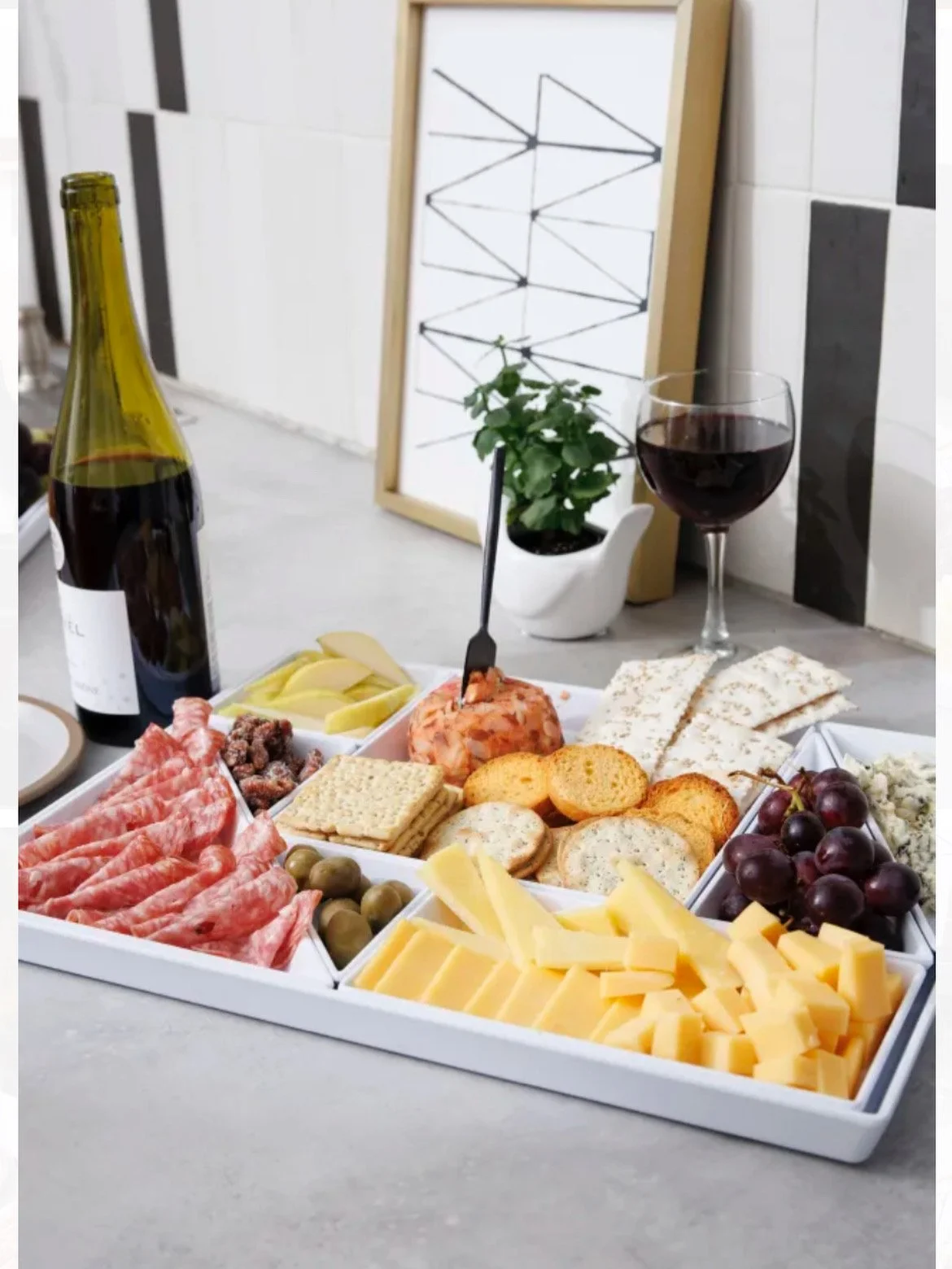 A cheese and charcuterie board with grapes, crackers, sliced cheese, cured meats, and spreads, accompanied by a glass of red wine and a bottle of wine on a kitchen counter.