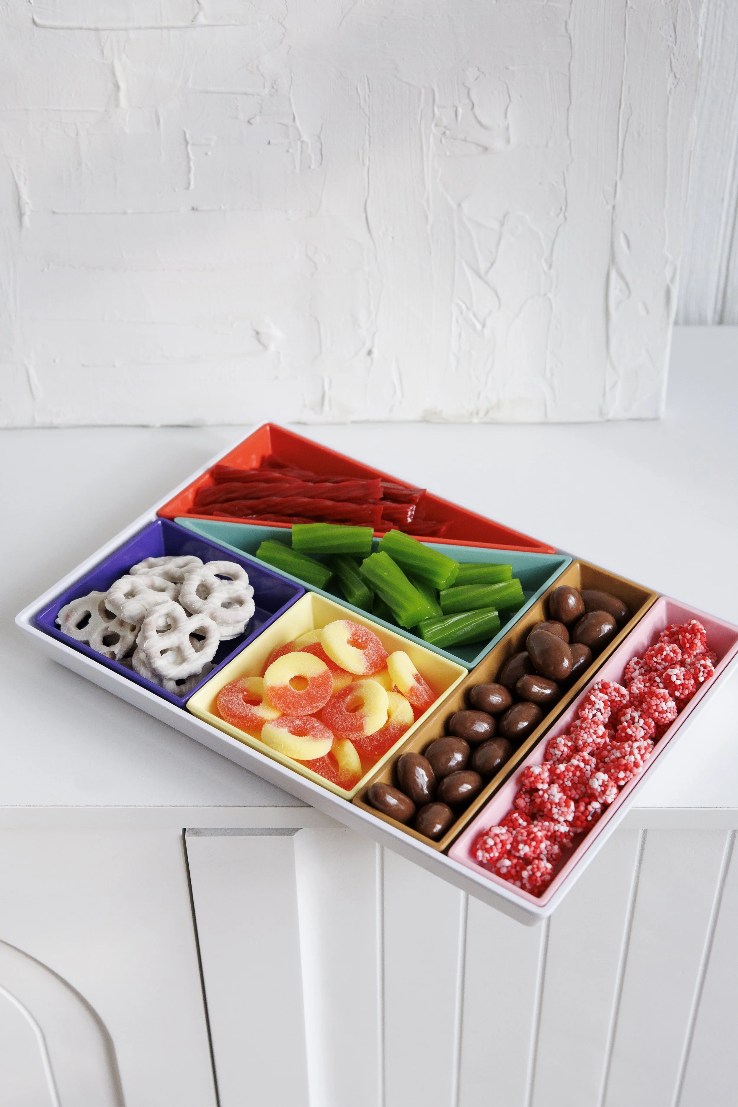 Colorful divided tray with assorted candies, including pretzels, green licorice, red and white gummy worms, chocolate-covered nuts, and decorated sugar-coated candies, on a white surface in front of a textured white wall.
