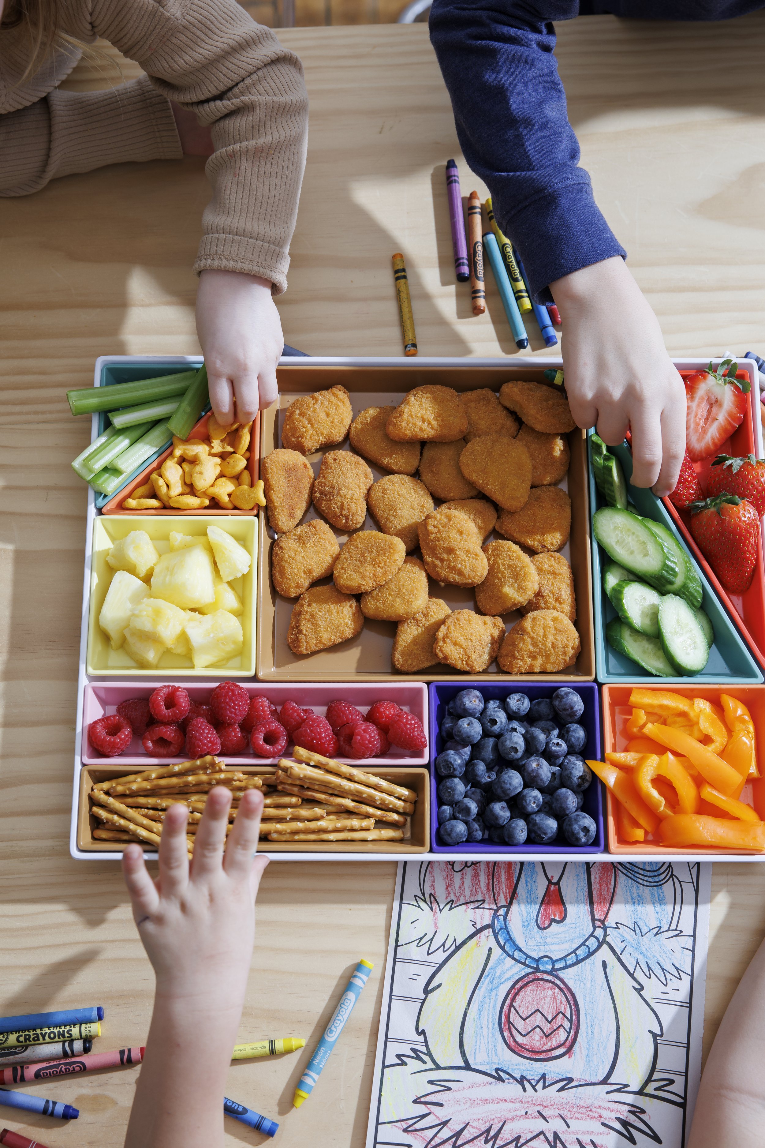 Children's hands reaching for food in a divided lunch box which contains chicken nuggets, celery sticks, pineapple chunks, strawberries, raspberries, blueberries, orange bell pepper slices, pretzel sticks, and a coloring page of a cartoon character.