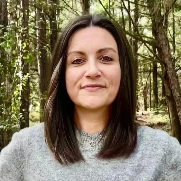A woman with shoulder-length dark hair standing outdoors in a wooded area.