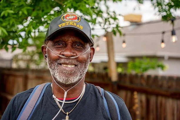 A smiling older man with a gray beard wearing a black cap with a military badge and 'VETERAN' embroidered on it, an oxygen tube, a navy blue shirt, and a backpack, standing outdoors with a wooden fence and string lights in the background.