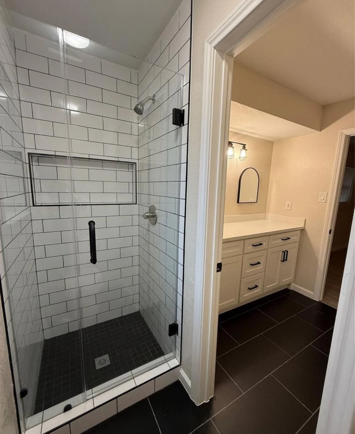 A bathroom with a glass-enclosed shower featuring white subway tiles and black grout; adjacent to a vanity with a white countertop and beige drawers, illuminated by a two-light fixture, with a mirror and wall outlets.