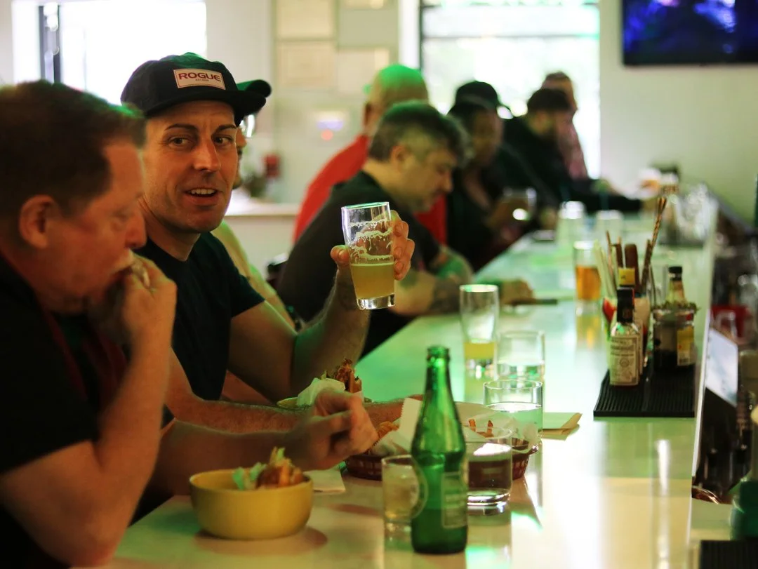 A group of people sitting at a bar counter enjoying drinks and food. One man is holding a beer and looking at the camera, wearing a black cap with a 'ROGUE' logo. Others are eating or drinking, with various beverages and condiments on the counter.