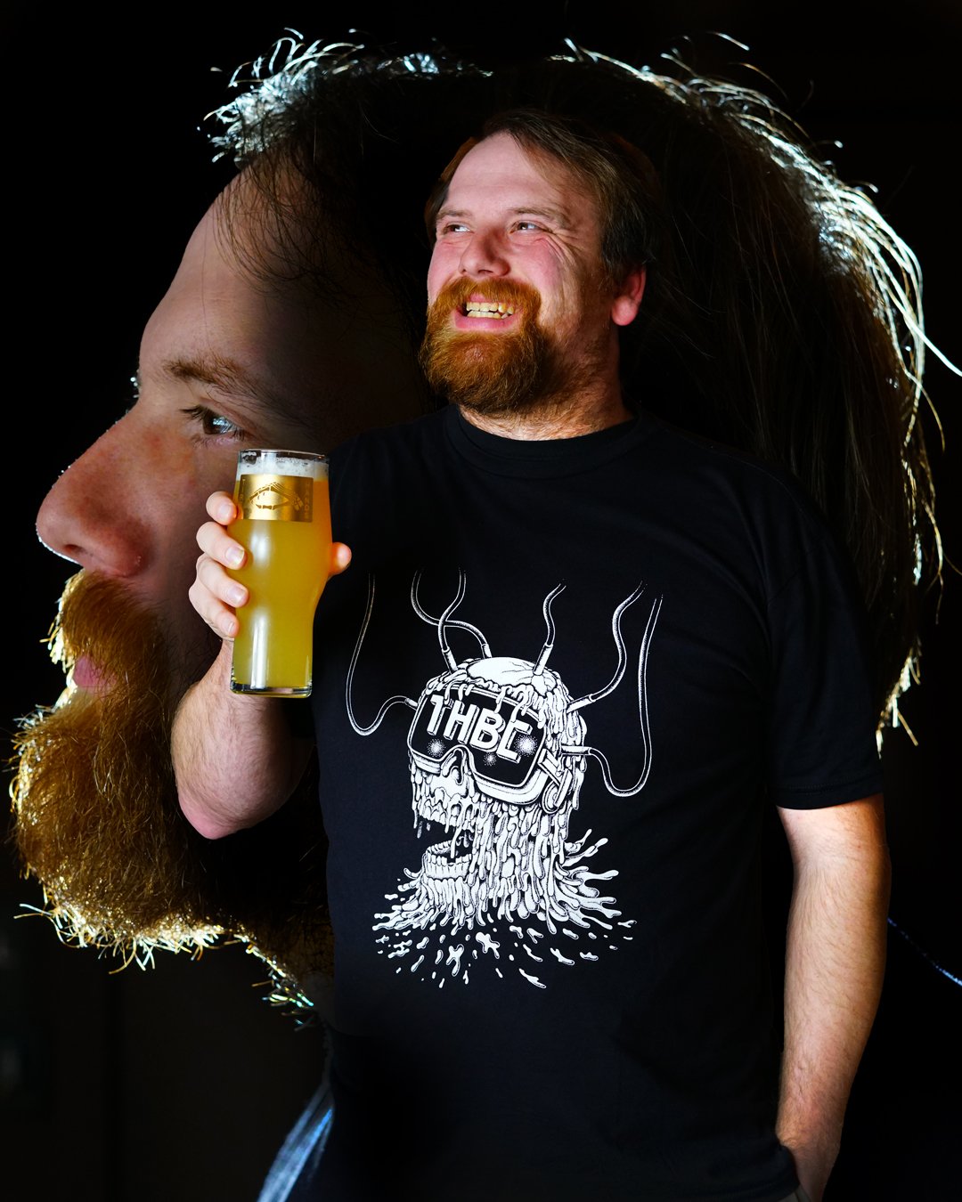 A man with a beard and long hair smiling, holding a glass of beer, standing in front of a large, semi-transparent image of his own face with long hair.