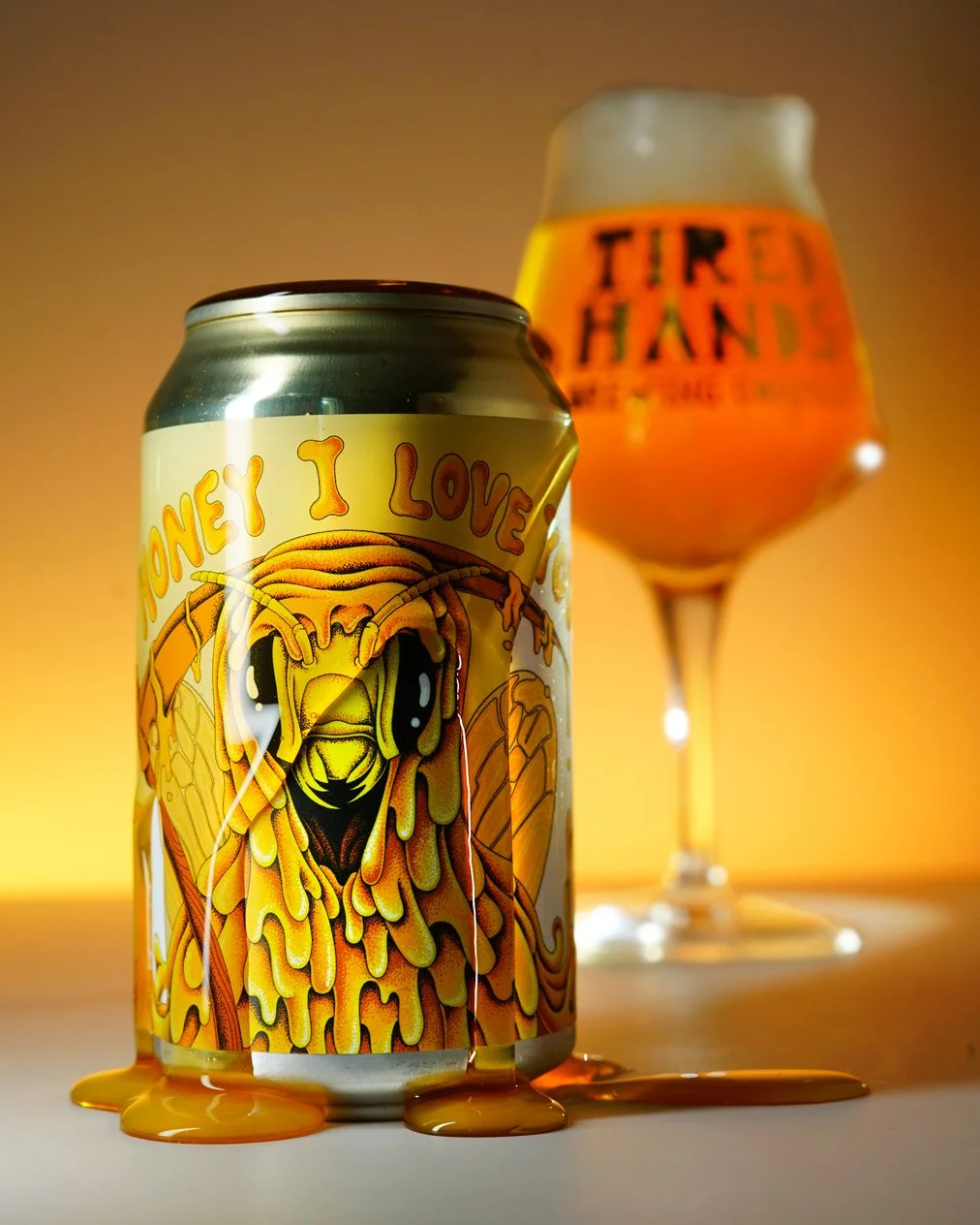 A can of beer with a colorful label featuring a bee and the words 'Honey I Love' and a glass of beer in the background with the words 'Tired Hands' on it, set against an orange and yellow background.