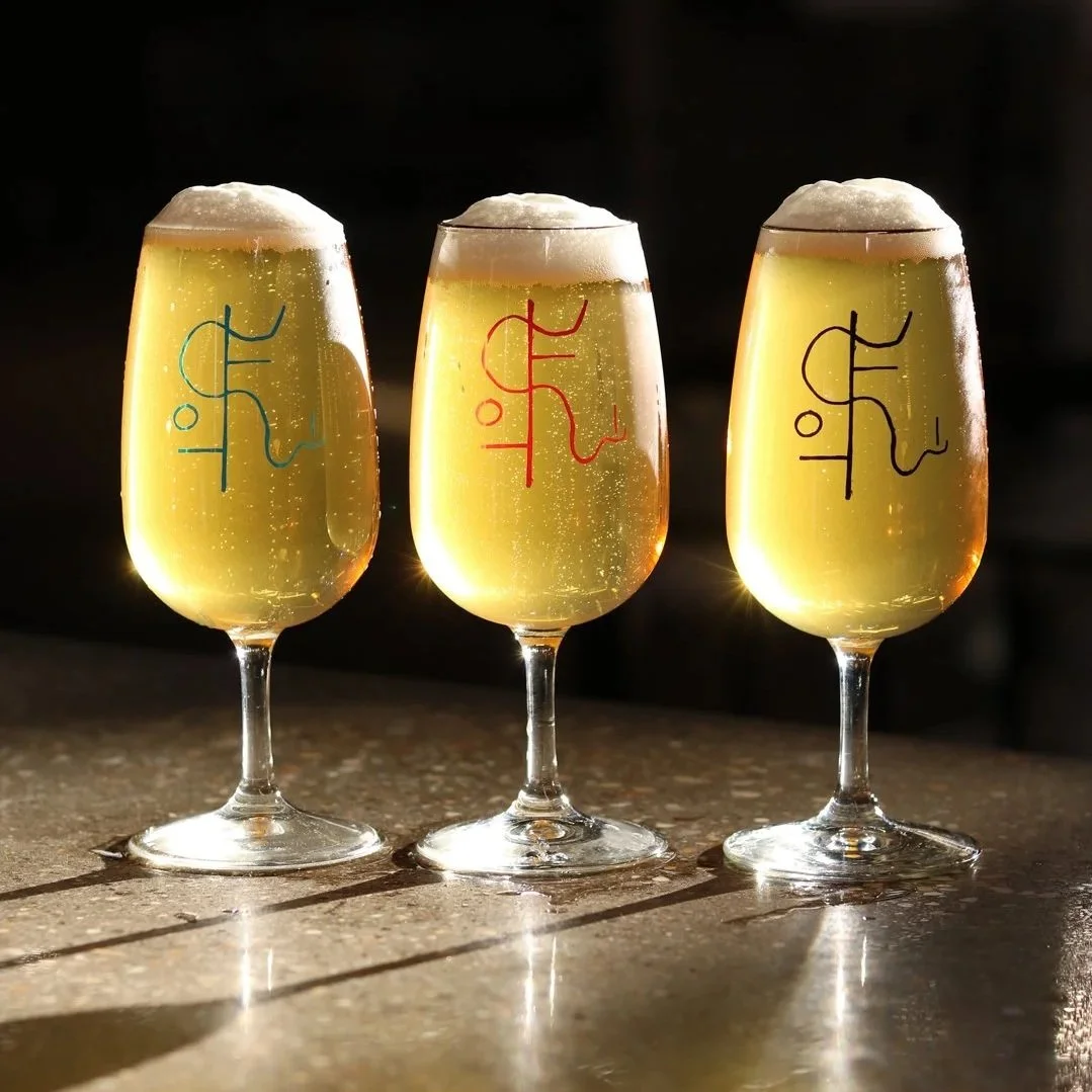 Three glasses of champagne with foam on top, each with a different colored logo on the front, placed on a surface with shadows cast by the glasses.