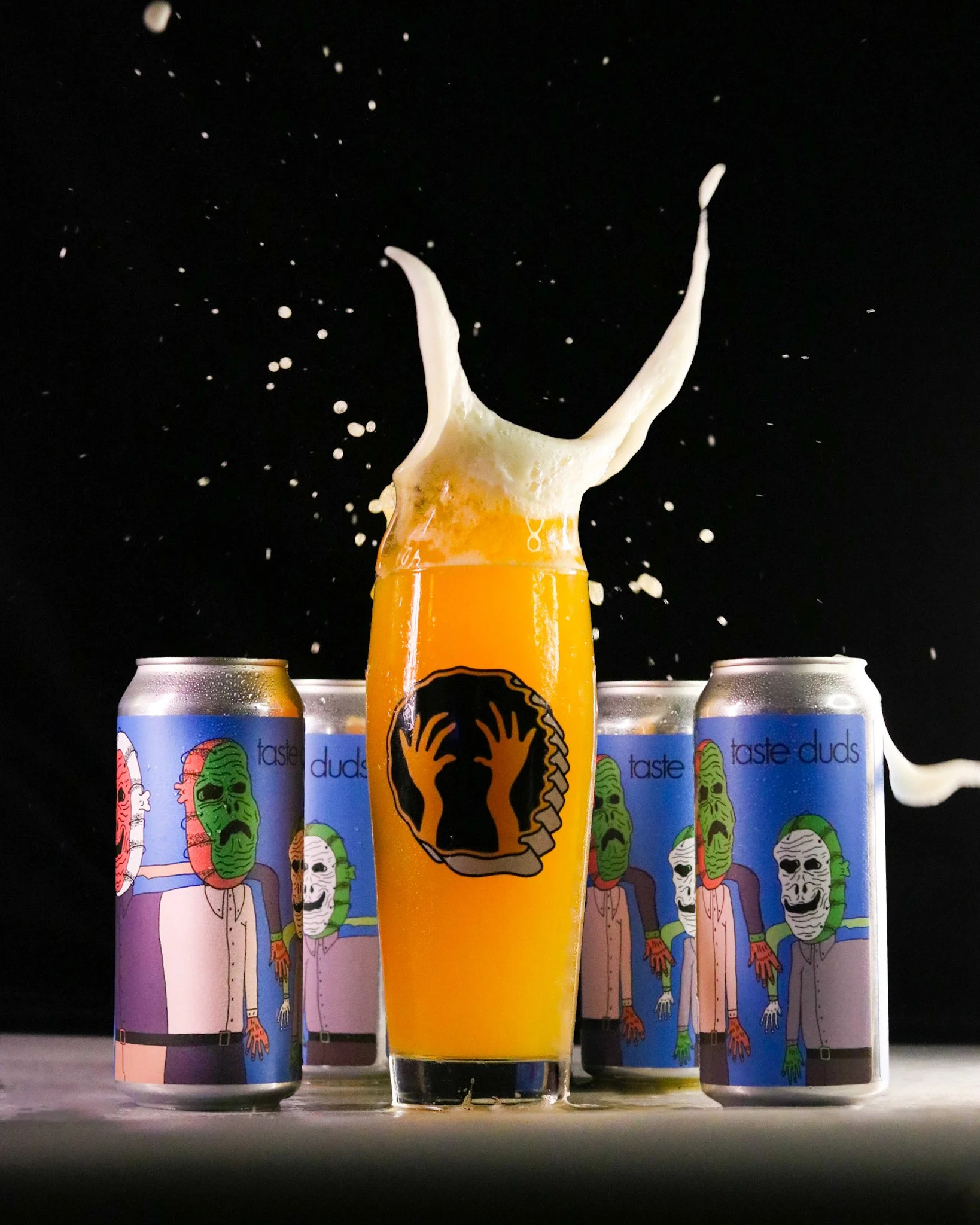 A beer glass overflowing with foam in front of four cans with cartoon characters wearing masks. The cans have the text 'taste duds' on them.