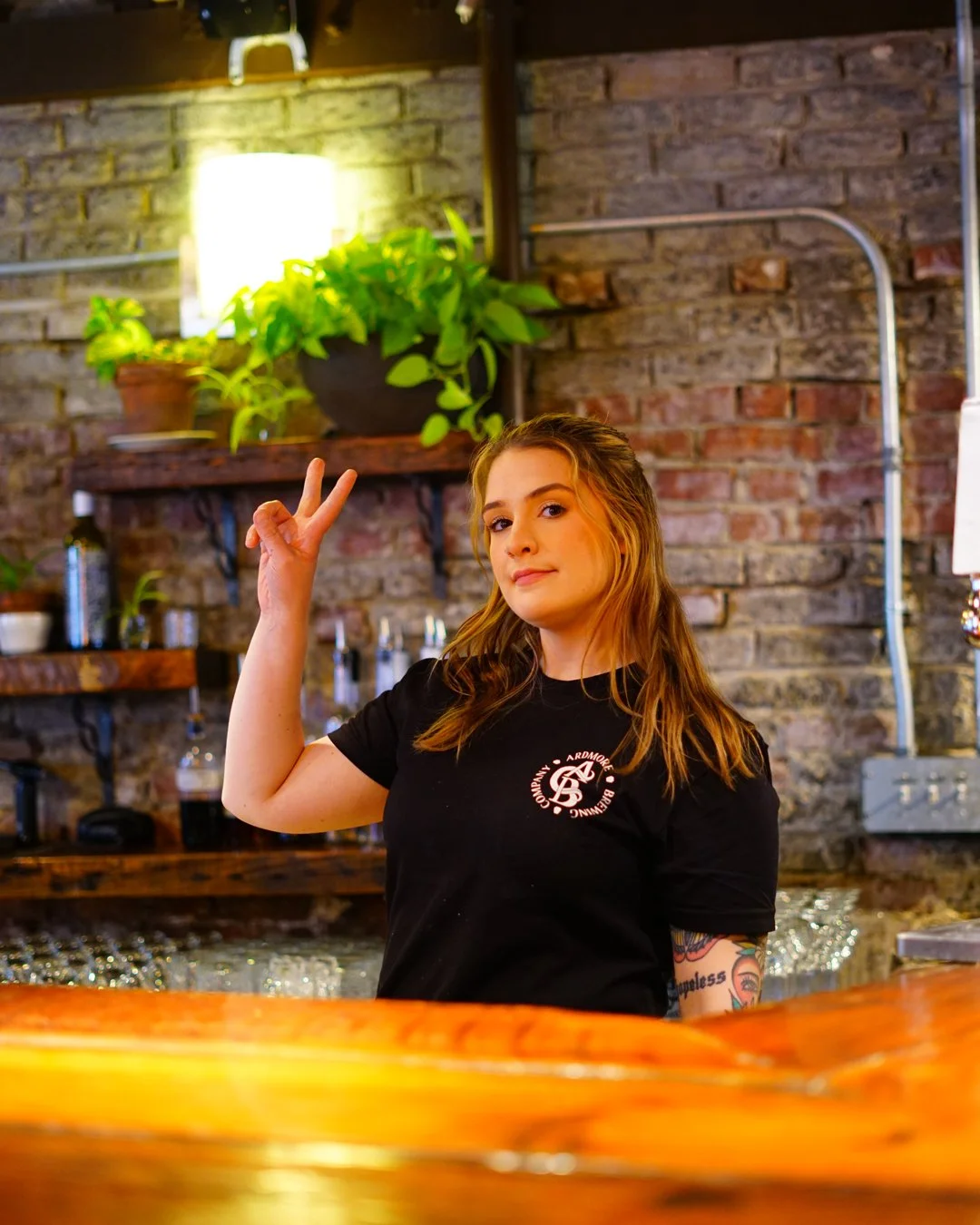 A young woman with wavy brown hair making a peace sign with her right hand inside a restaurant or bar. She is wearing a black T-shirt with a logo and has visible tattoos on her left arm. The background features a brick wall, wooden shelves with green potted plants, a bright lamp, and various bar accessories.