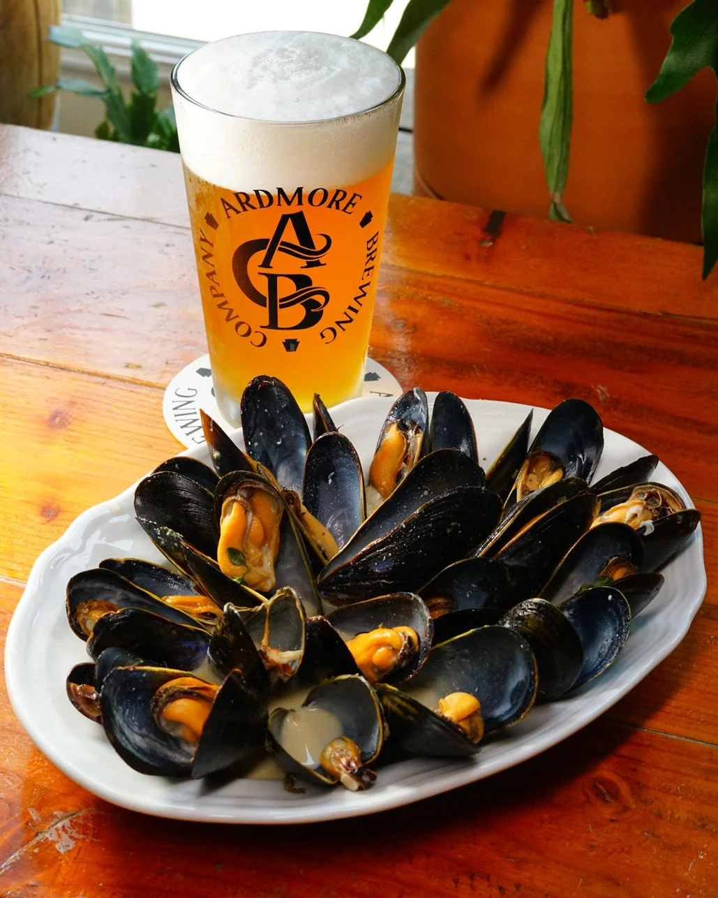 A white plate of cooked mussels with orange flesh, a glass of beer with foam, and a wooden table with a plant in the background.