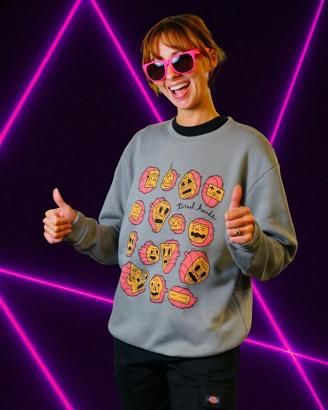 Woman wearing sunglasses and a gray sweatshirt with cartoon faces and the words "tired hands", giving thumbs up in front of a black background with pink neon lines.