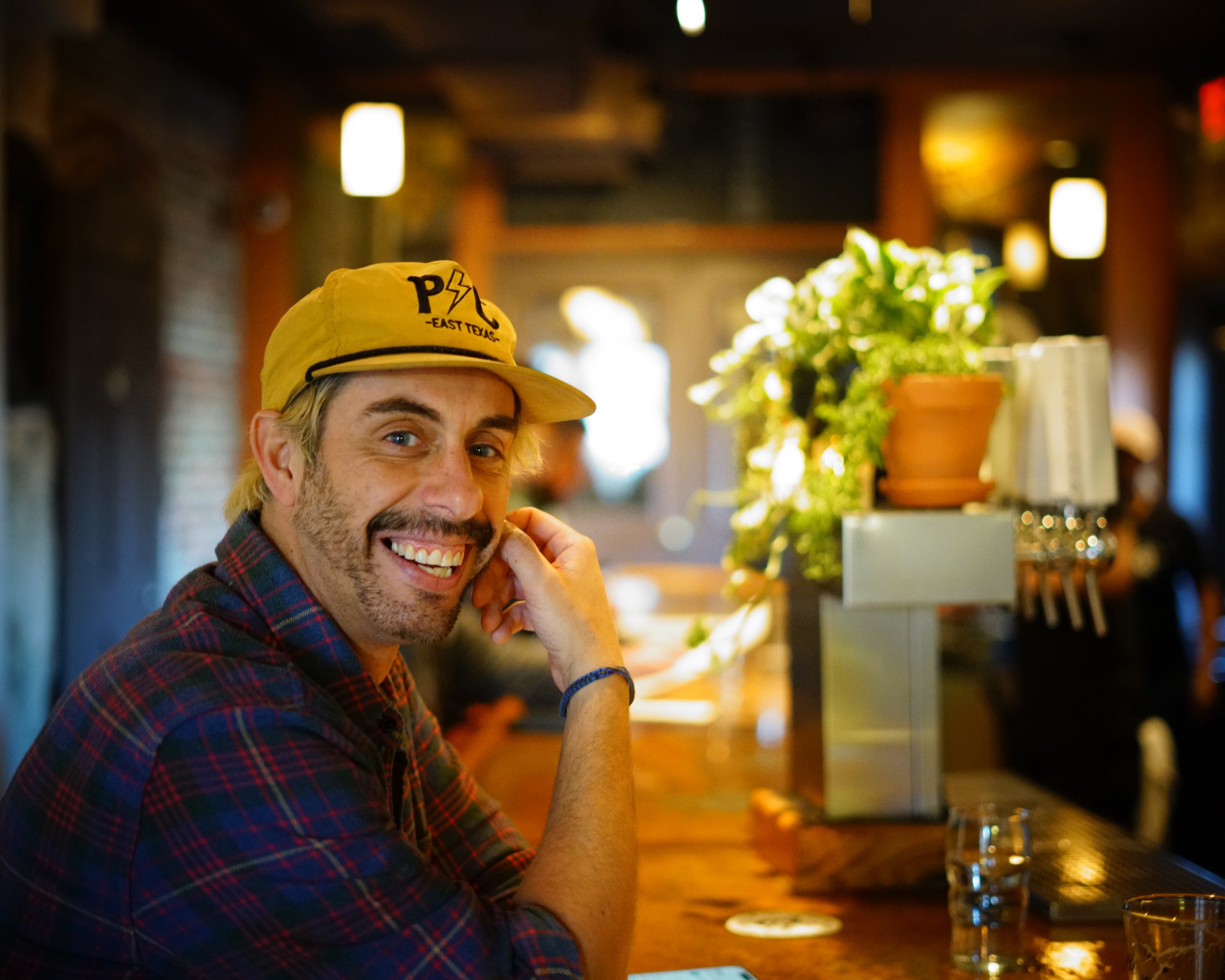 A man with a beard and a yellow cap smiling at the camera while sitting at a bar.