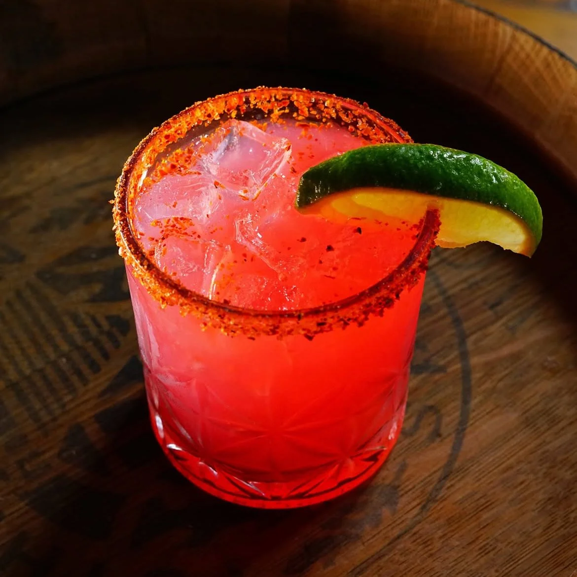 A pink cocktail in a textured glass with a salted rim, garnished with a lime wedge, served on a wooden surface.