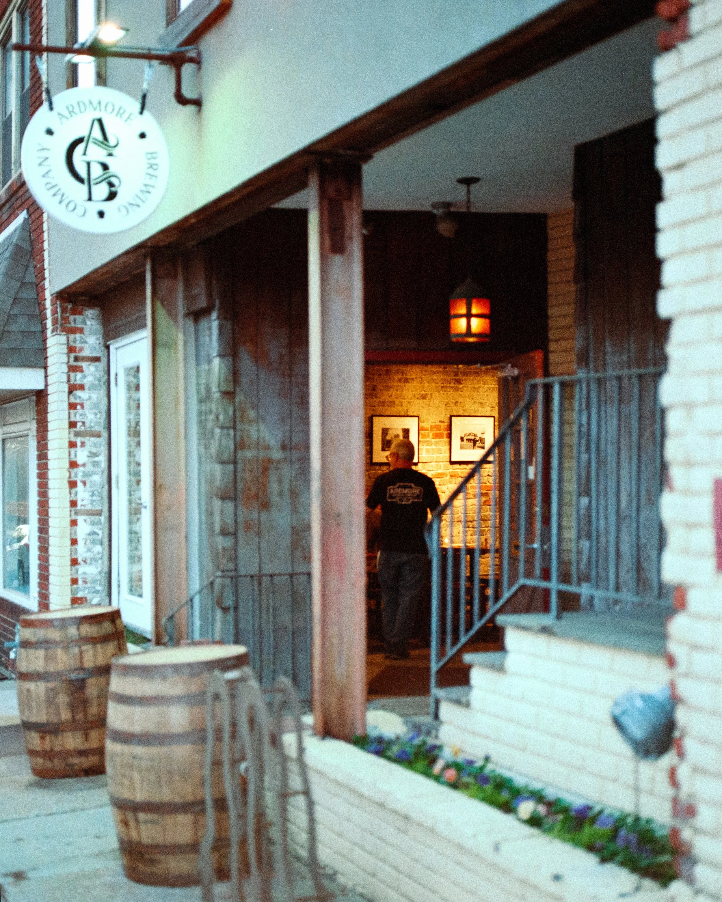 Exterior of a building with a hanging sign for Ardmore Brewing Company, showing barrels and outdoor seating. Inside, a person is standing near framed photographs on a brick wall.