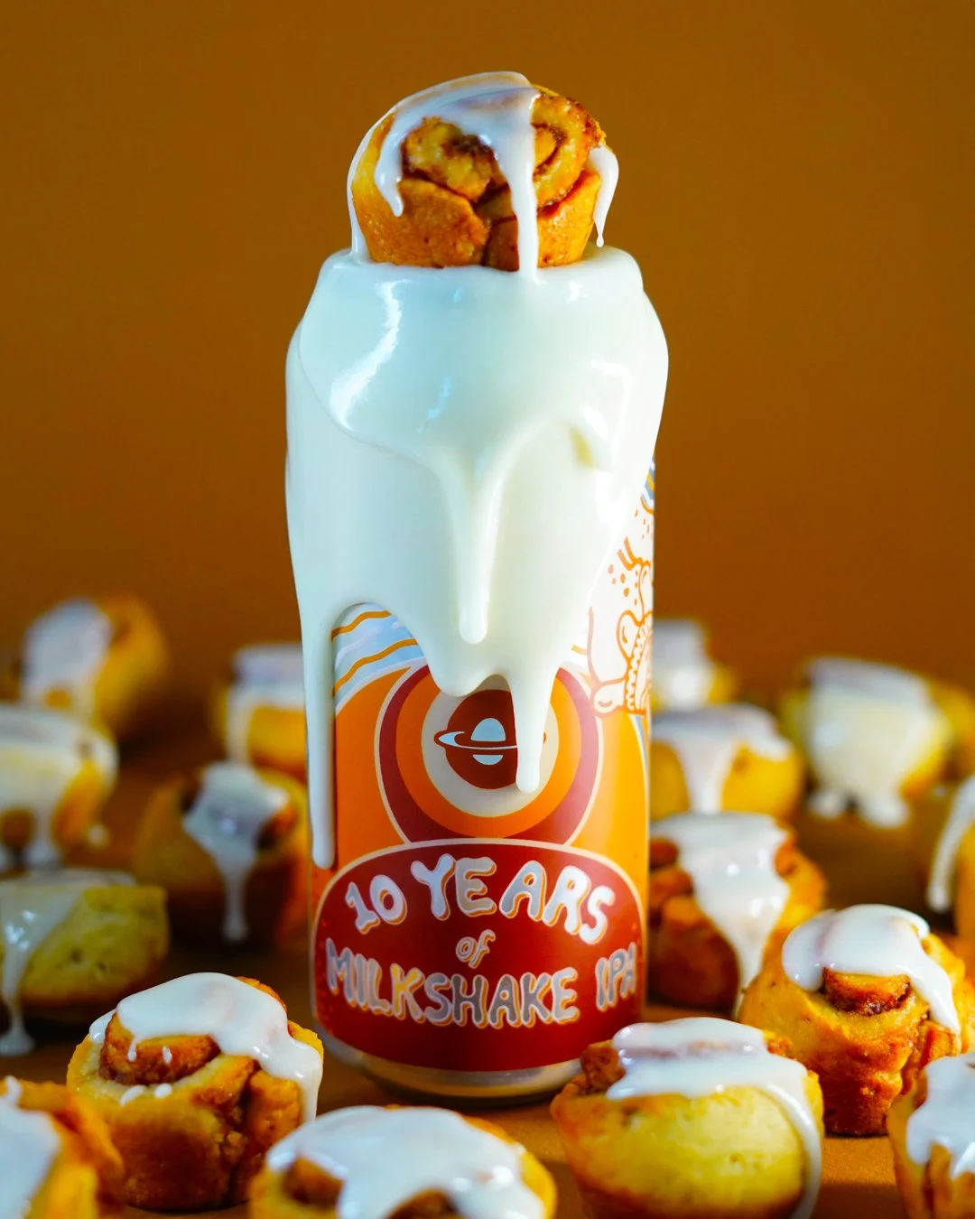 A can of milkshake IPA beer with melted cream overflowing on top, surrounded by mini muffins with icing.