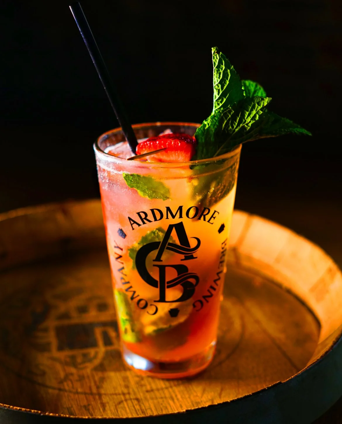 A colorful cocktail in a tall glass garnished with a strawberry, mint leaves, and a black straw, with the logo 'Ardmore' on the glass, placed on a wooden tray.