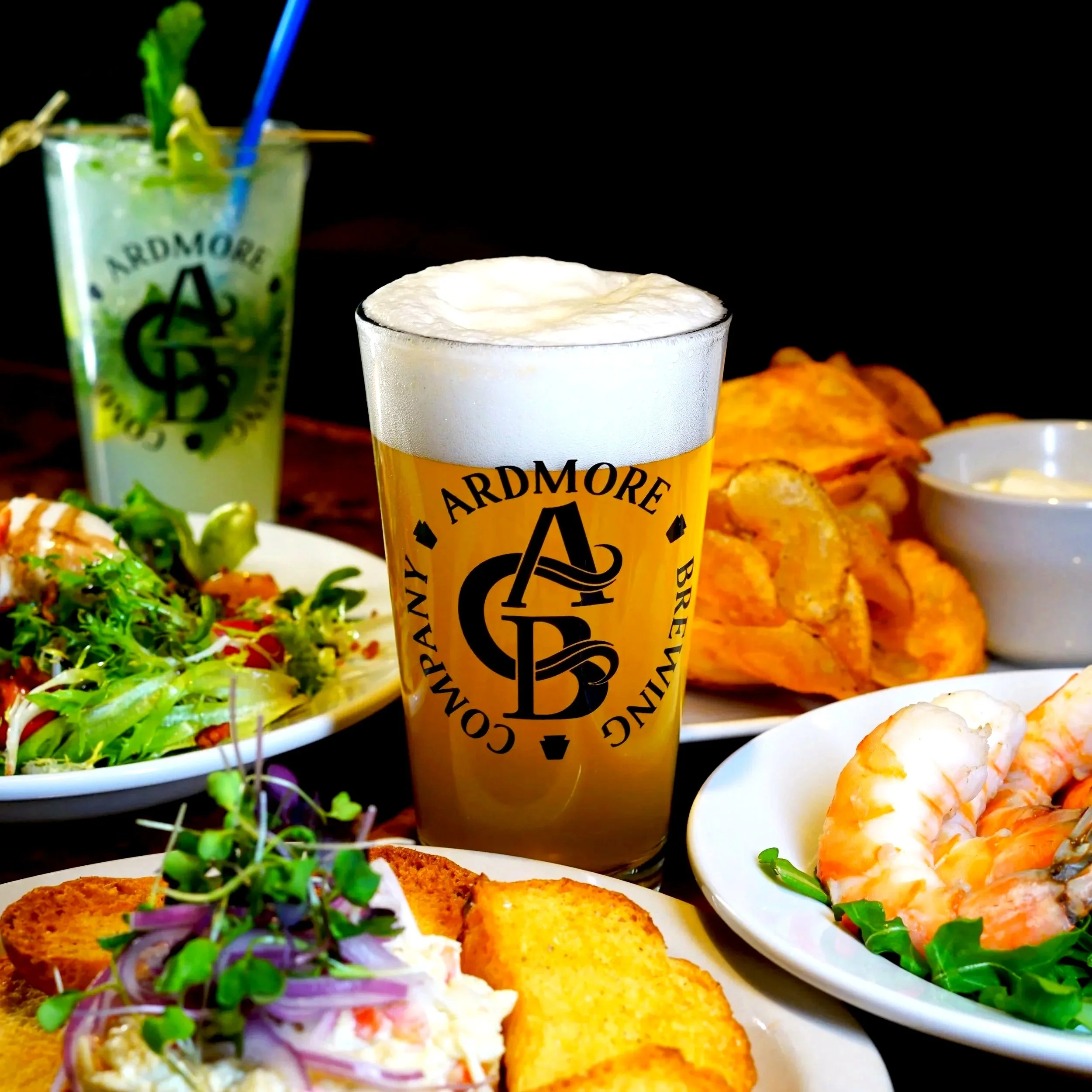 A table with a glass of pale beer topped with foam, a plate of fried shrimp, a salad, and a drink with lime and mint garnish.