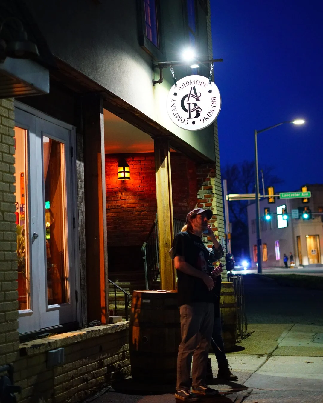 People standing outside a brewery at night, talking on a mobile phone, with a wooden barrel and brick wall in the background. The brewery sign is visible above and street lights illuminate the scene.