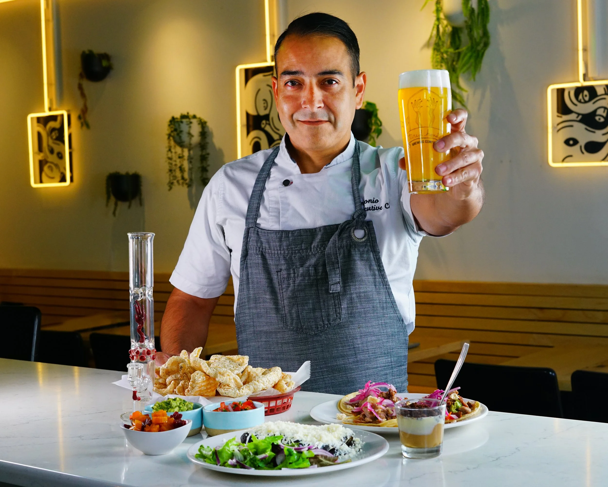 A chef in a white uniform and gray apron is holding a glass of beer in a restaurant. The counter in front of him has various dishes including salads, breaded food, and a layered dessert. The background has decorative plants and modern art lighting.