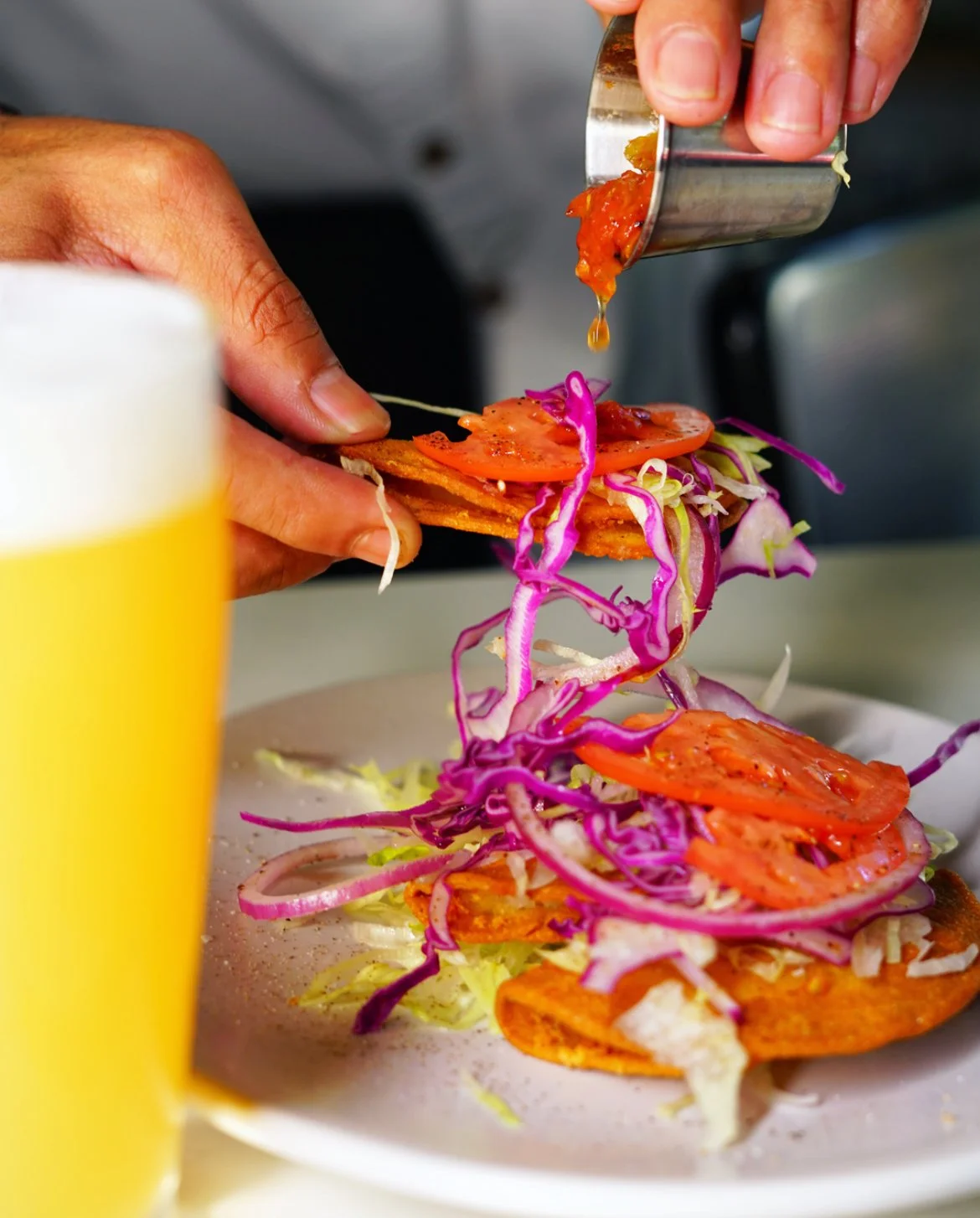 Person pouring sauce onto a taco with sliced tomatoes, shredded cabbage, and crispy shells, with a glass of yellow drink in foreground.
