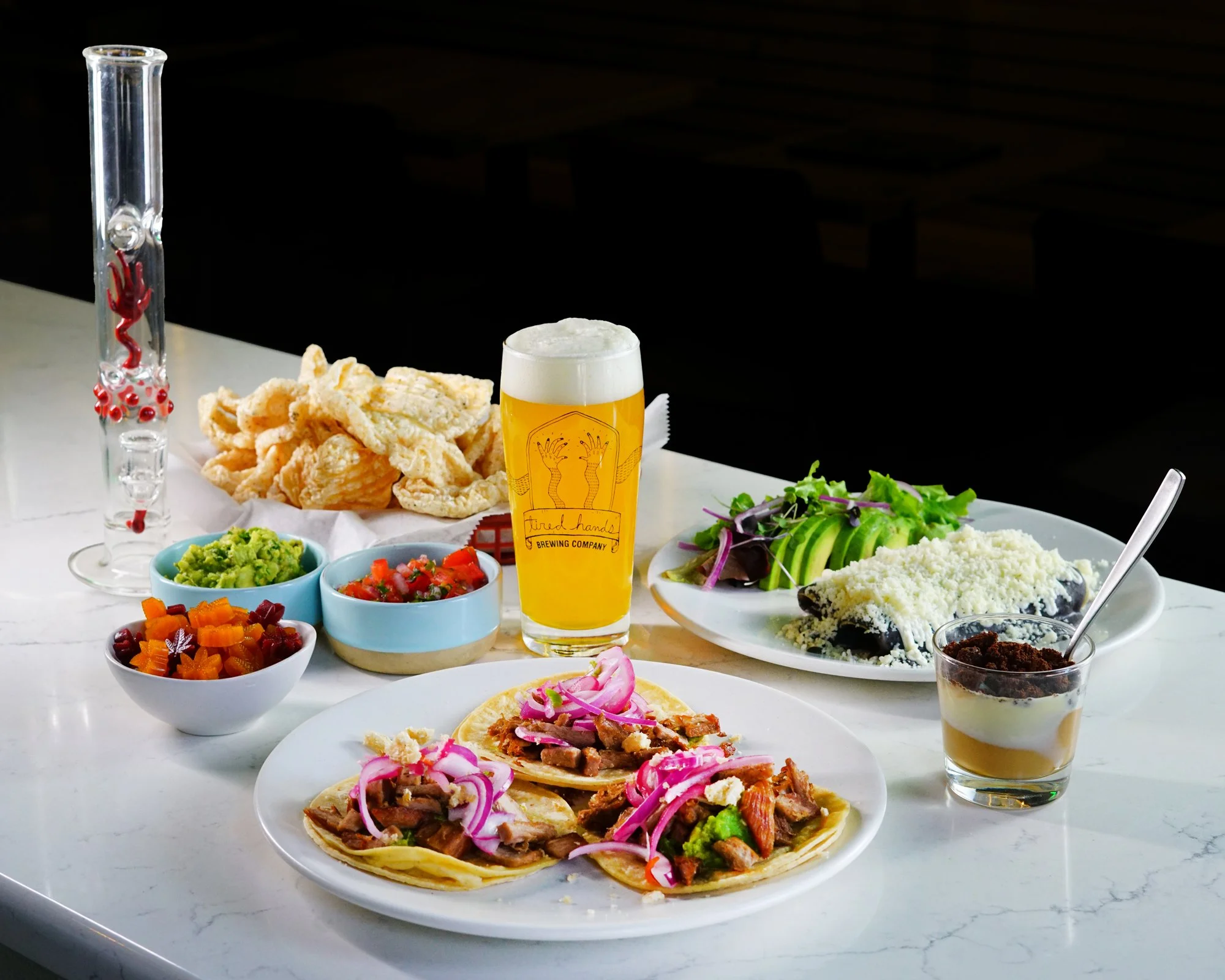 Assorted Mexican dishes and drinks on a white marble table, including tacos, pickled vegetables, guacamole, salsa, a plate of pastel de queso, a glass of beer, and a glass of layered dessert, with a white background.
