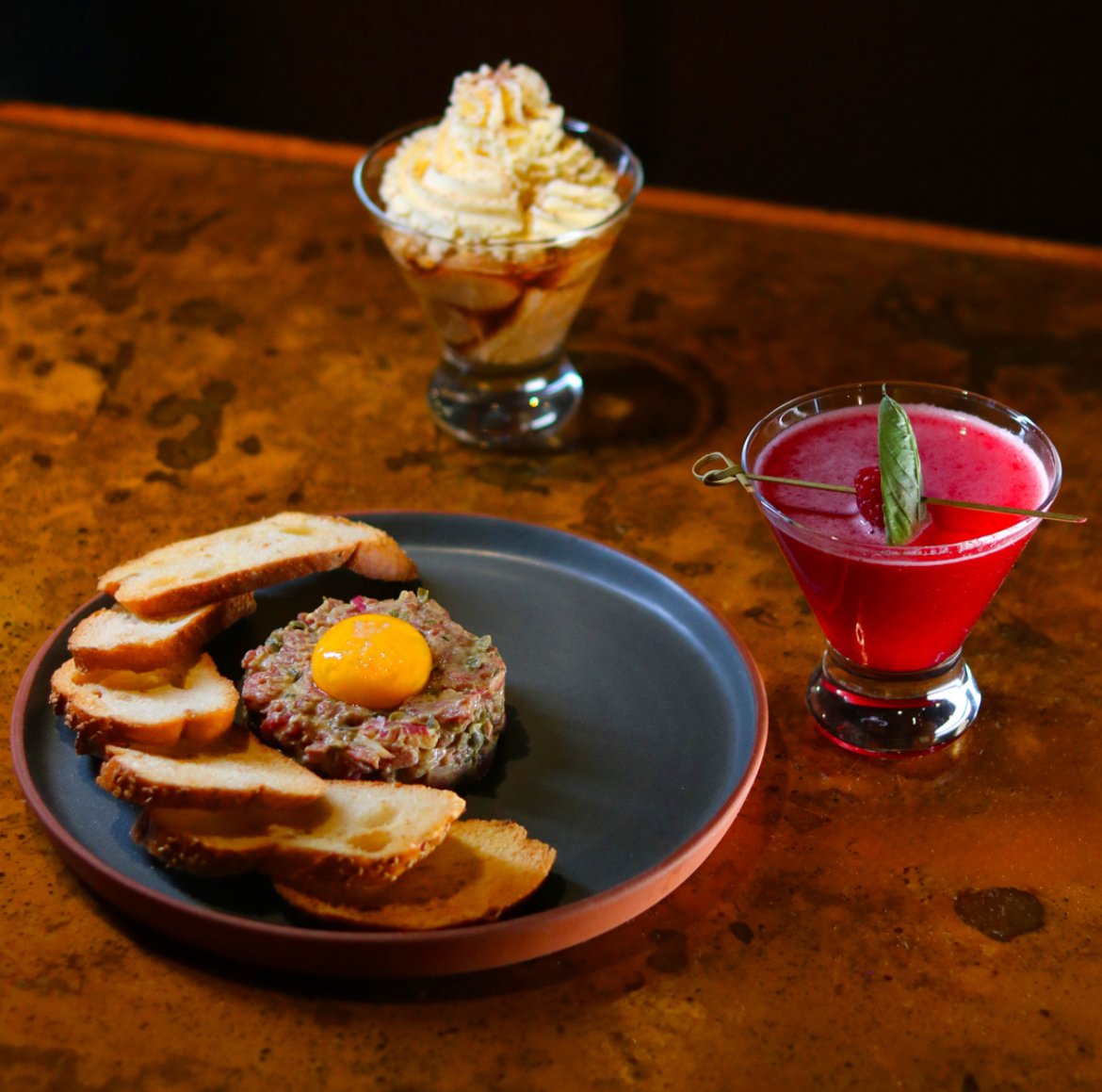 A plate with bread slices and steak tartare topped with a raw egg yolk, two cocktails (one red with a cherry and leaf garnish, and one with whipped cream and caramel drizzle) on a wooden table.