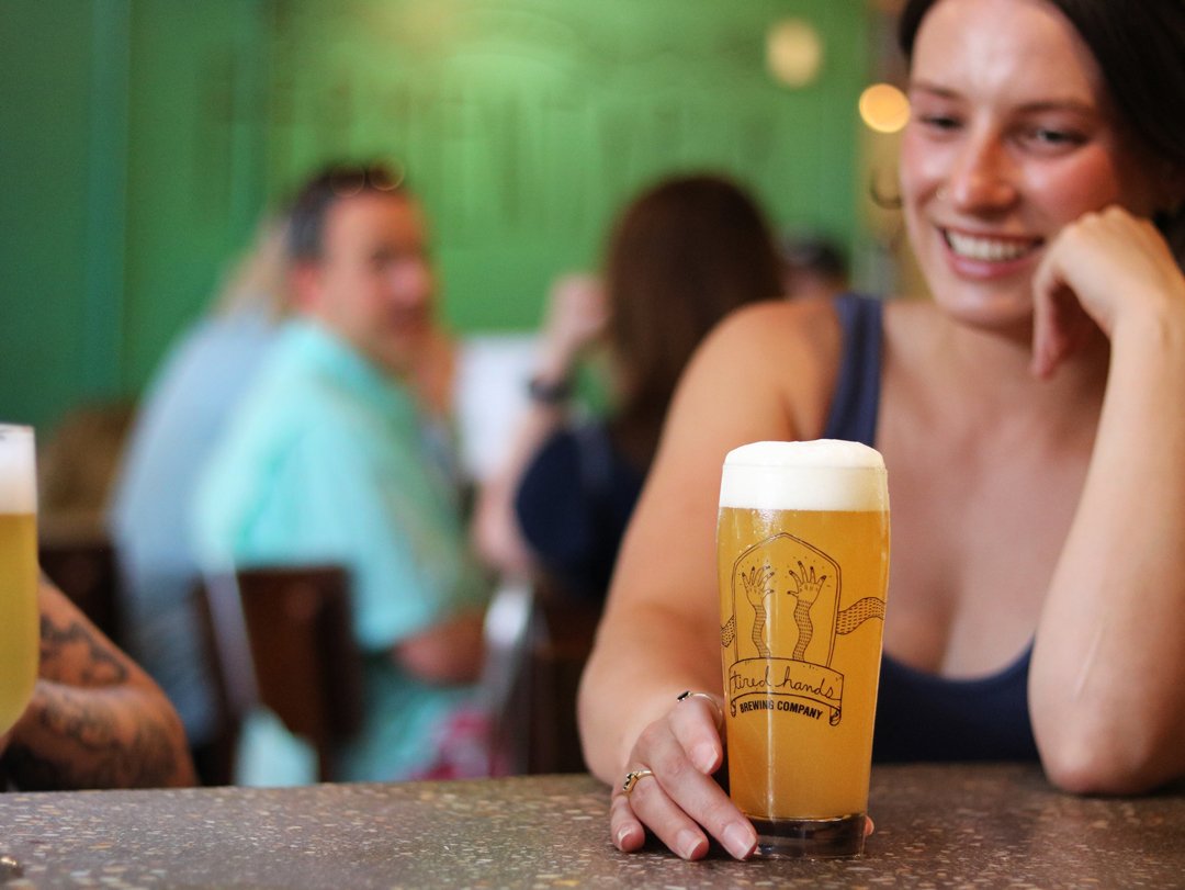 A woman smiling while sitting at a bar with a glass of beer in front of her. The beer glass has a logo that says 'Fired Hands Brewing Company.' Other people are blurred in the background.