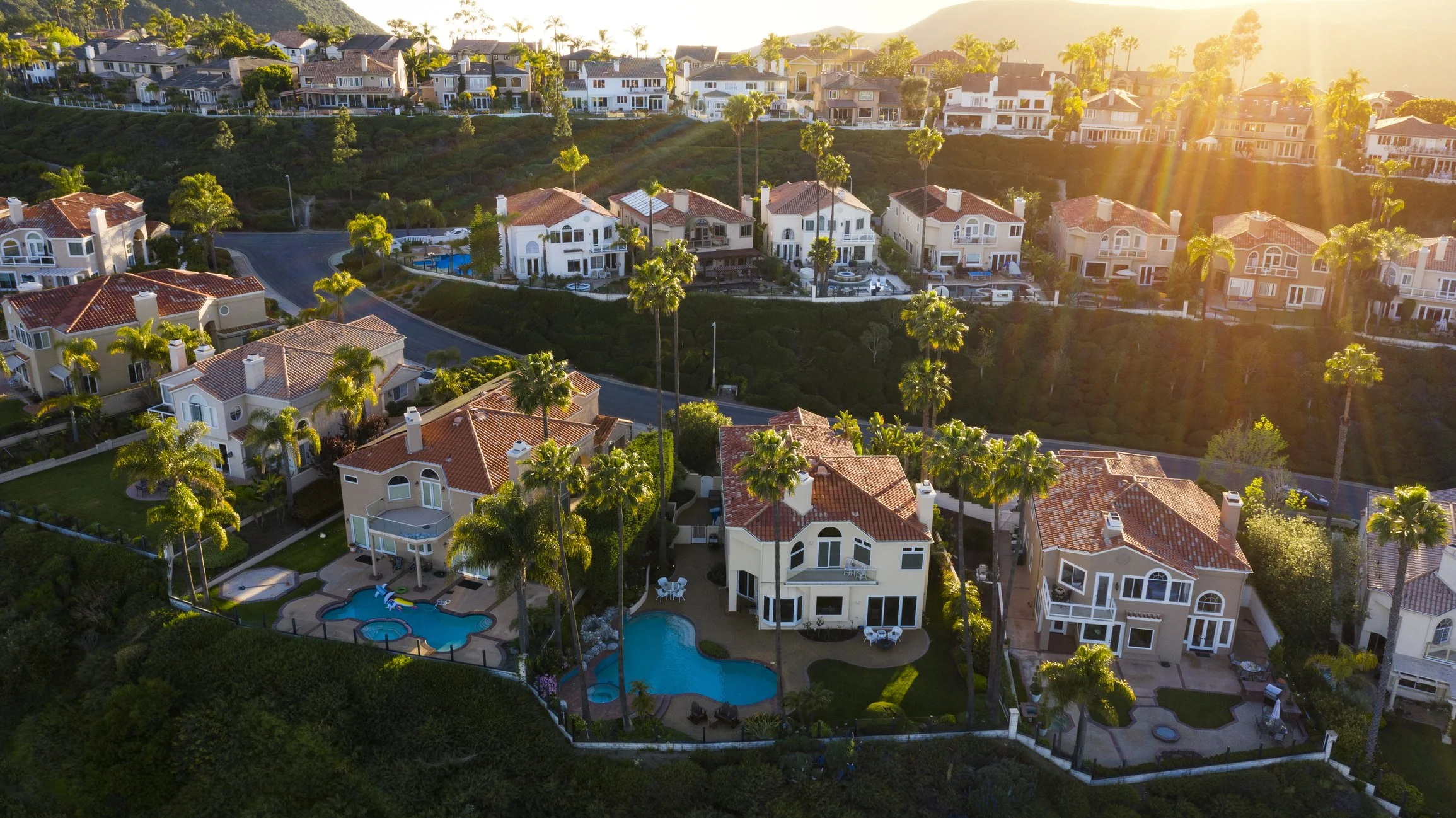 Aerial view of a hillside residential neighborhood with houses that have red tile roofs, palm trees, and swimming pools in backyards during sunset.