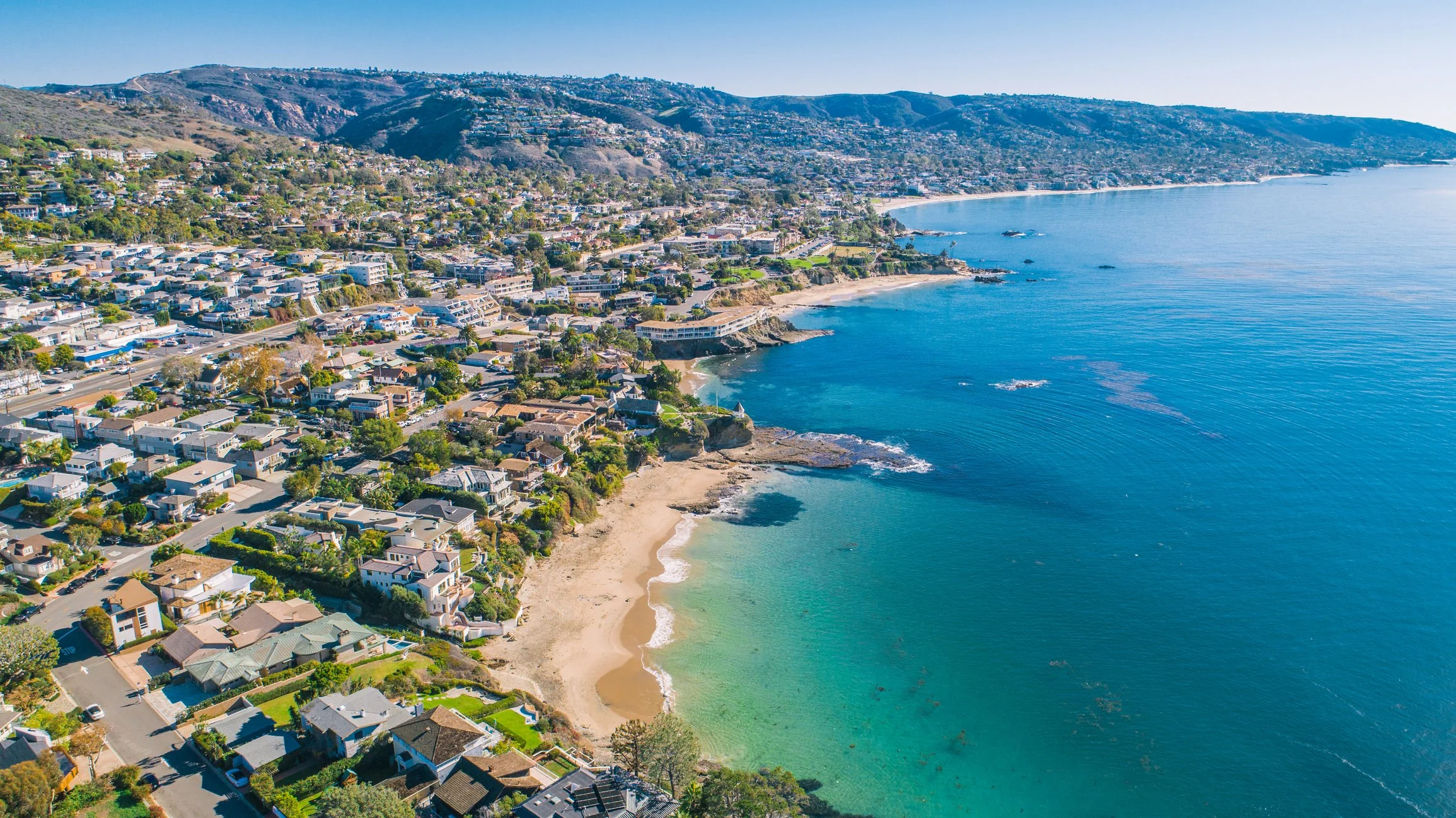 Aerial view of a coastal residential neighborhood with houses along the shoreline, sandy beaches, and hillside terrain, adjacent to the calm blue ocean.
