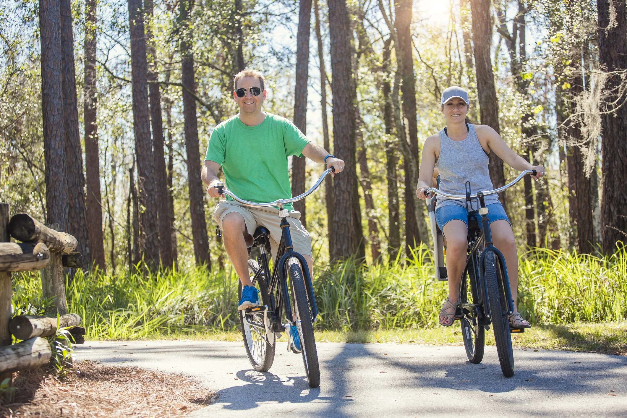 A man and woman riding bikes on a forest trail during daytime, both smiling and wearing casual summer clothing.